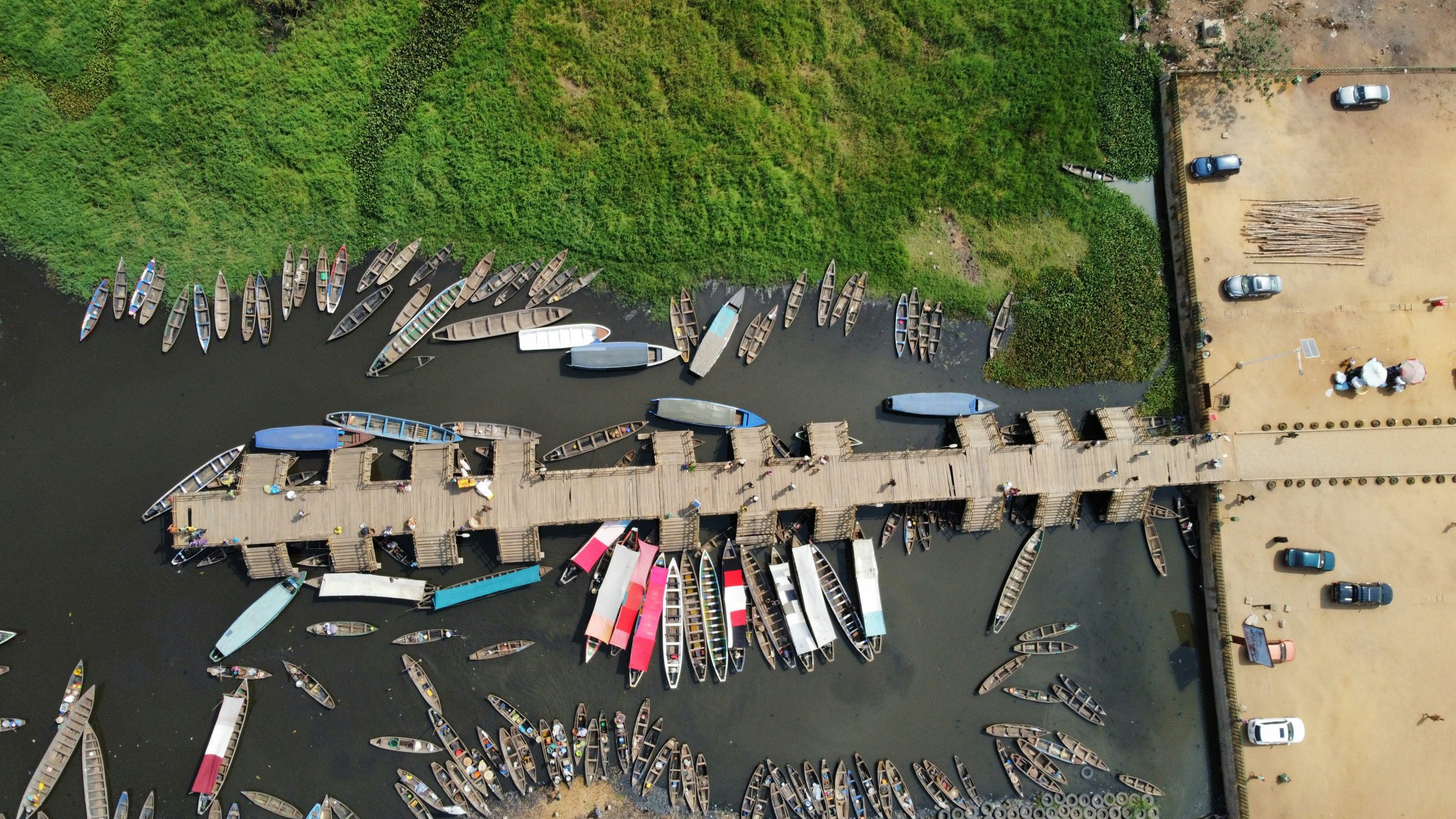 An overhead view of a bustling pier in Benin with numerous wooden canoes docked along the shore, surrounded by green vegetation.
