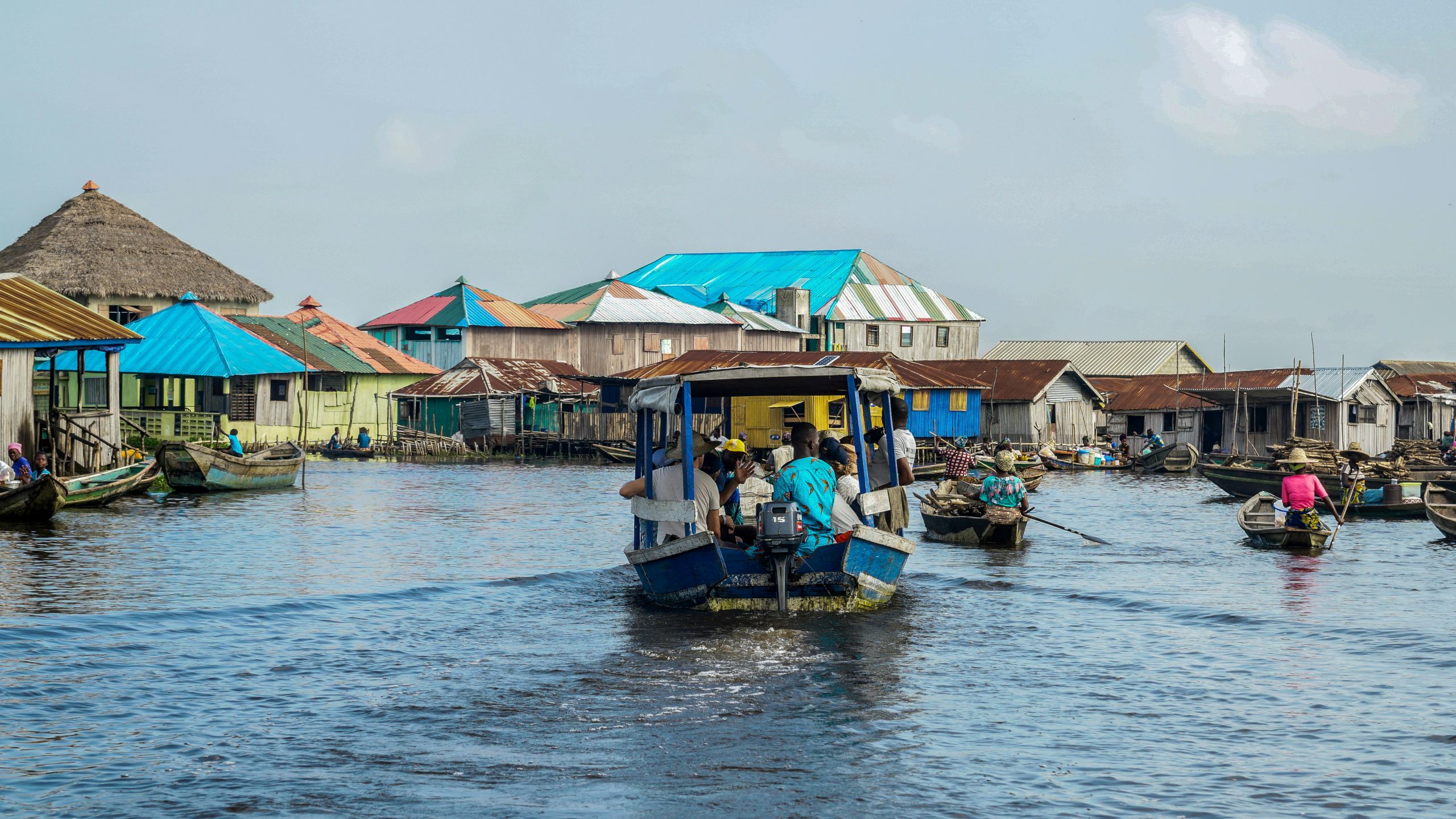 A vibrant scene of a water village in Benin with people navigating canoes, colorful rooftops, and a boat carrying passengers, illustrating daily life and local transportation.