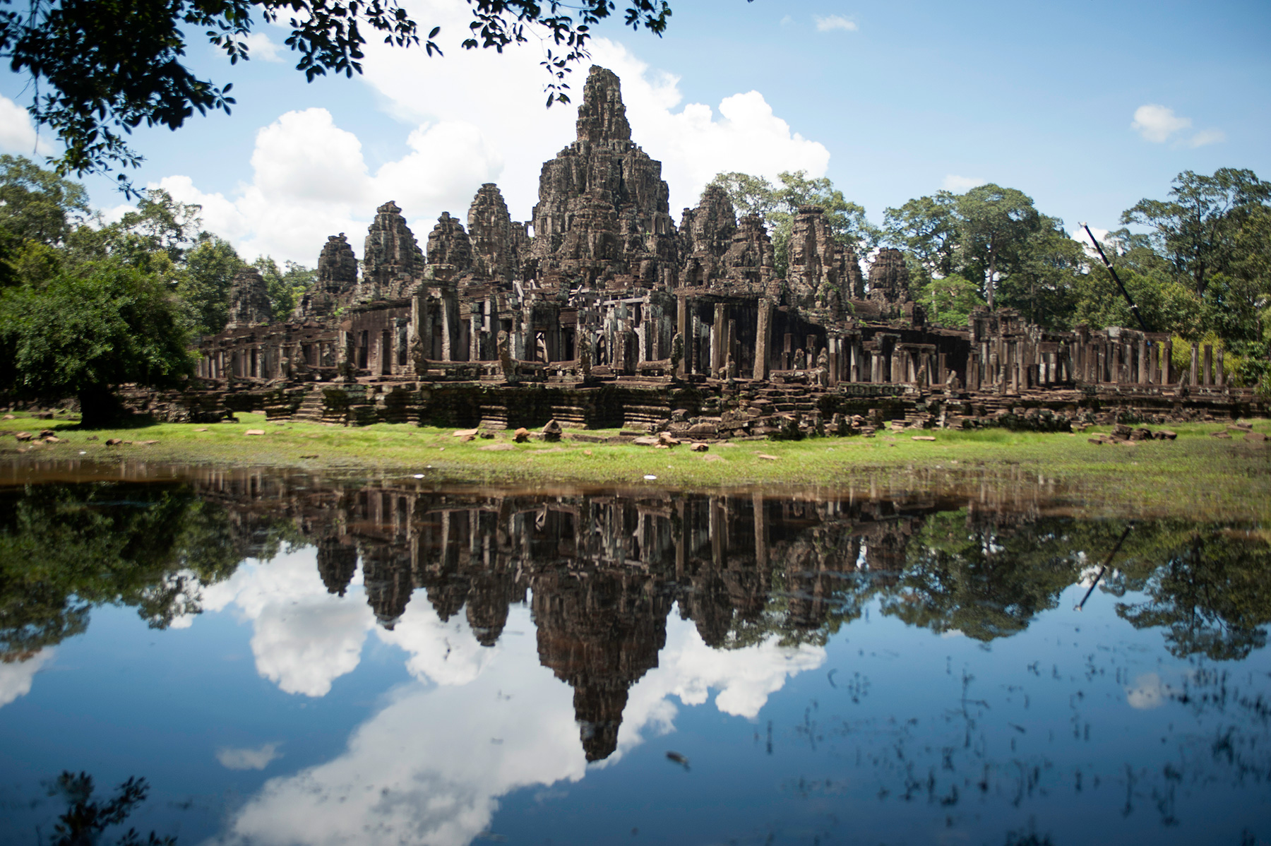 The face towers of the Bayon temple at Angkor Thom, mirrored in still water in the foreground under a blue sky.