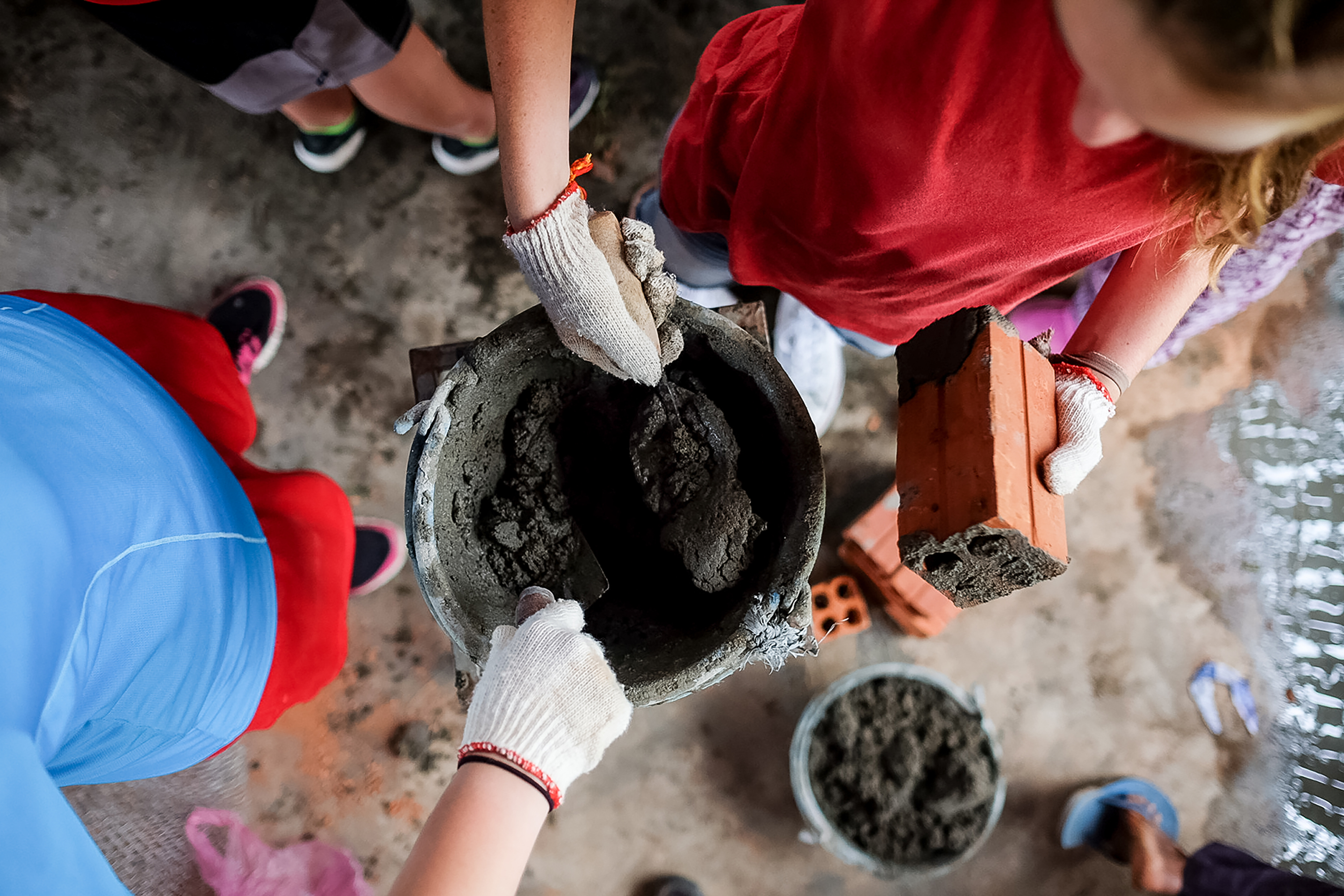 Students mix wet cement by hand near stacked bricks at a construction site