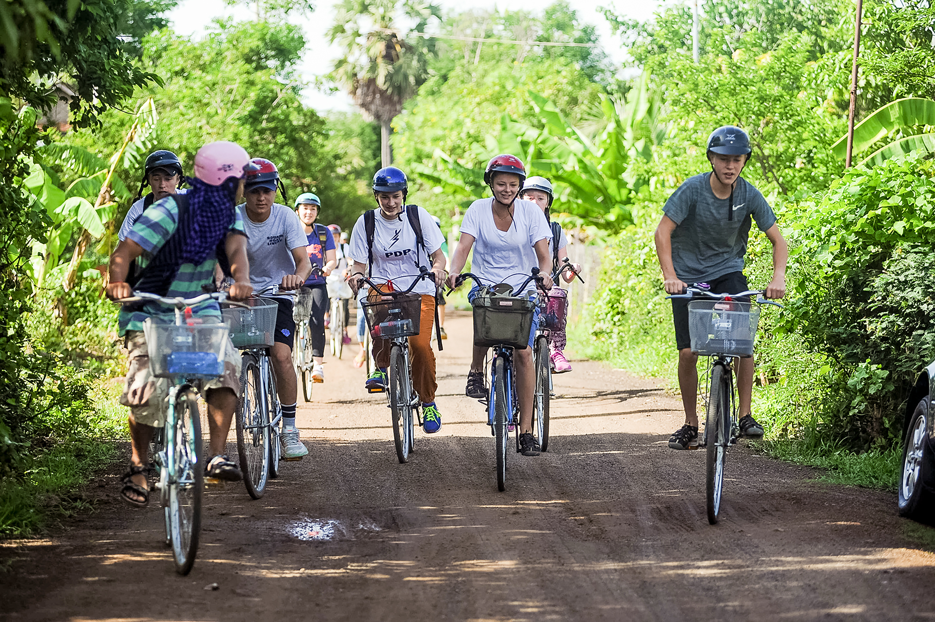 Students ride bicycles with wire baskets along a dirt road lined with palms