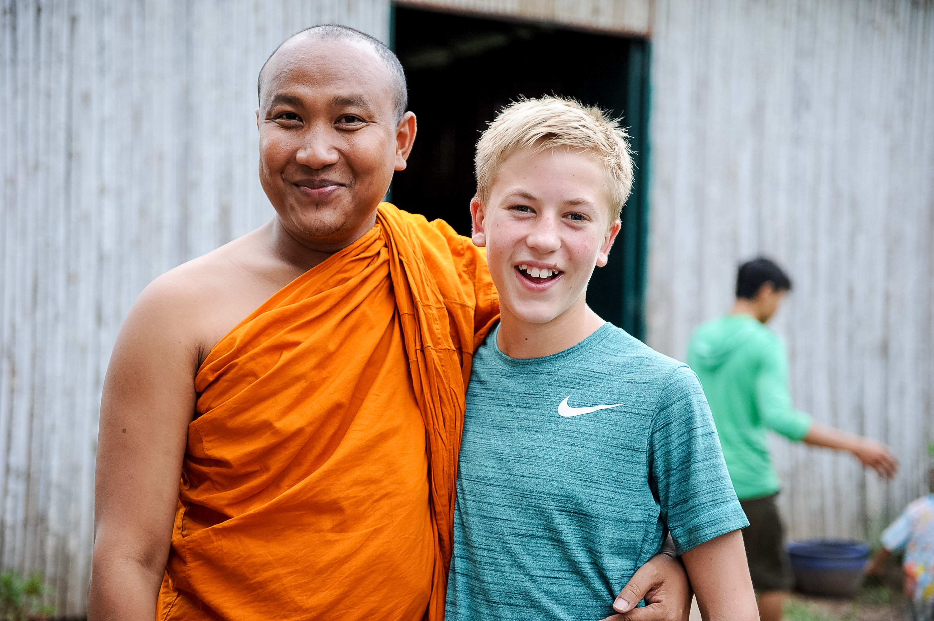 A student poses with a Buddhist monk in an orange robe outside a wooden building