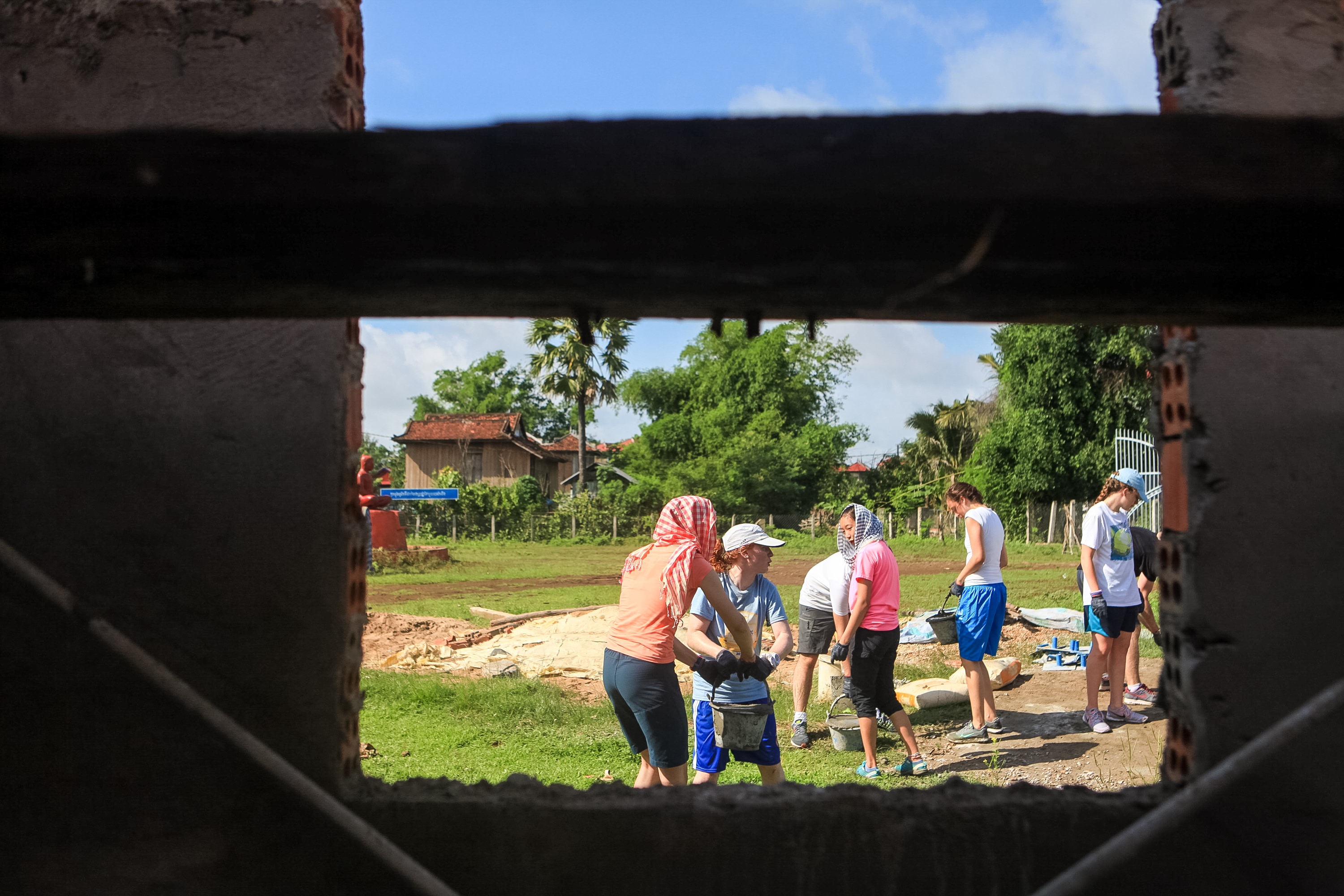 Seen through an unfinished window opening, students carry buckets and mix concrete on a village school building site in Kampong Cham Province.