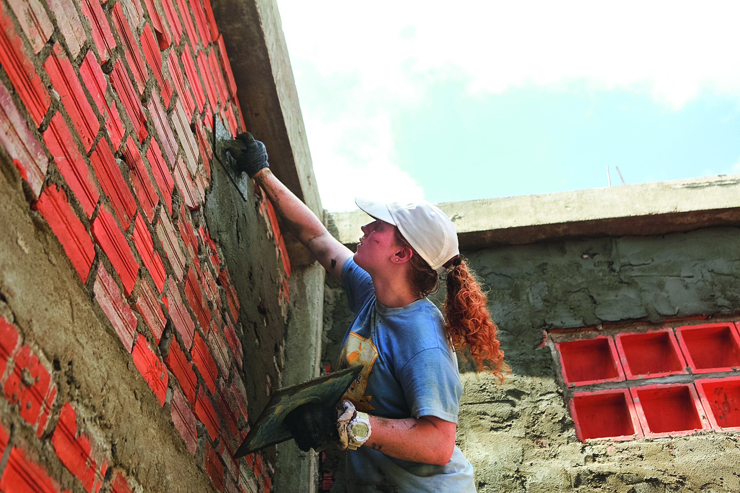 A student in a white cap renders a brick wall with a trowel and float on a school building site in Kampong Cham Province.
