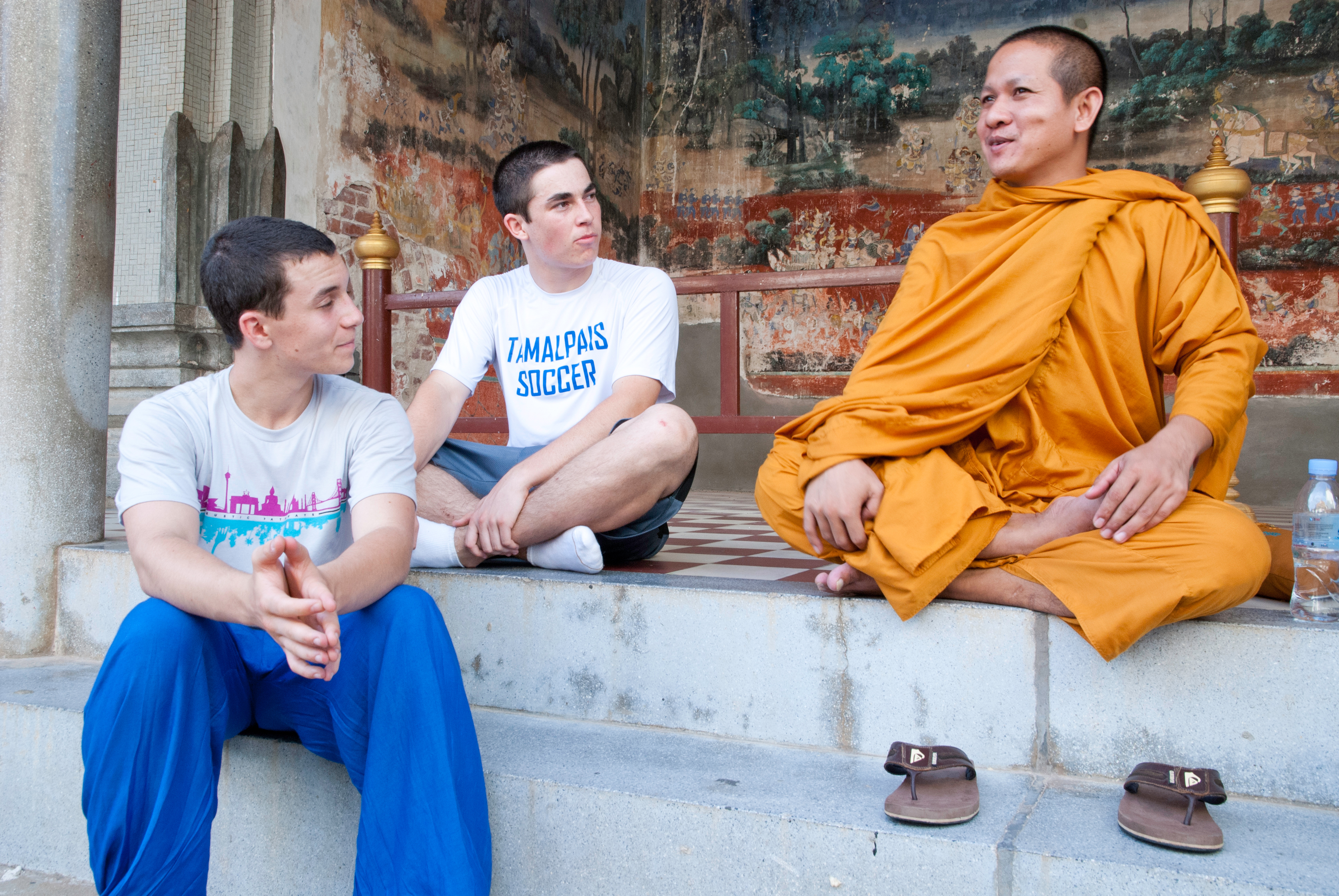 Two students sit on a temple step beside a monk in orange robes, with painted murals on the wall behind them, in Phnom Penh.