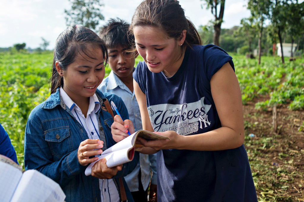 A student writes in a notebook with a local resident in a field