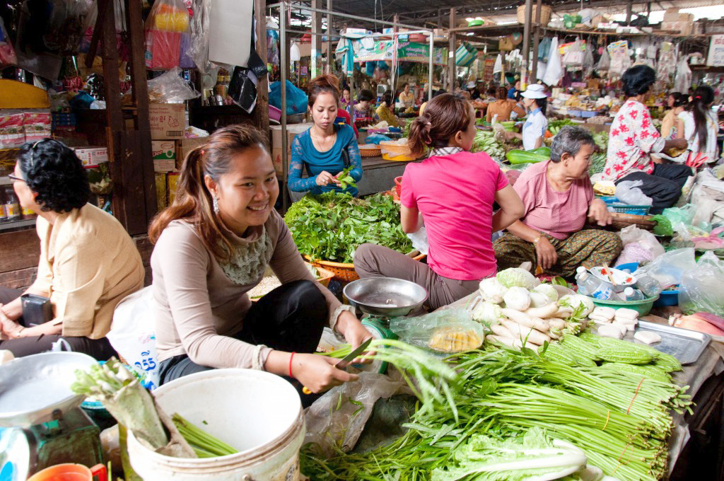 Students browse a covered market stall as vendors sort fresh greens and vegetables