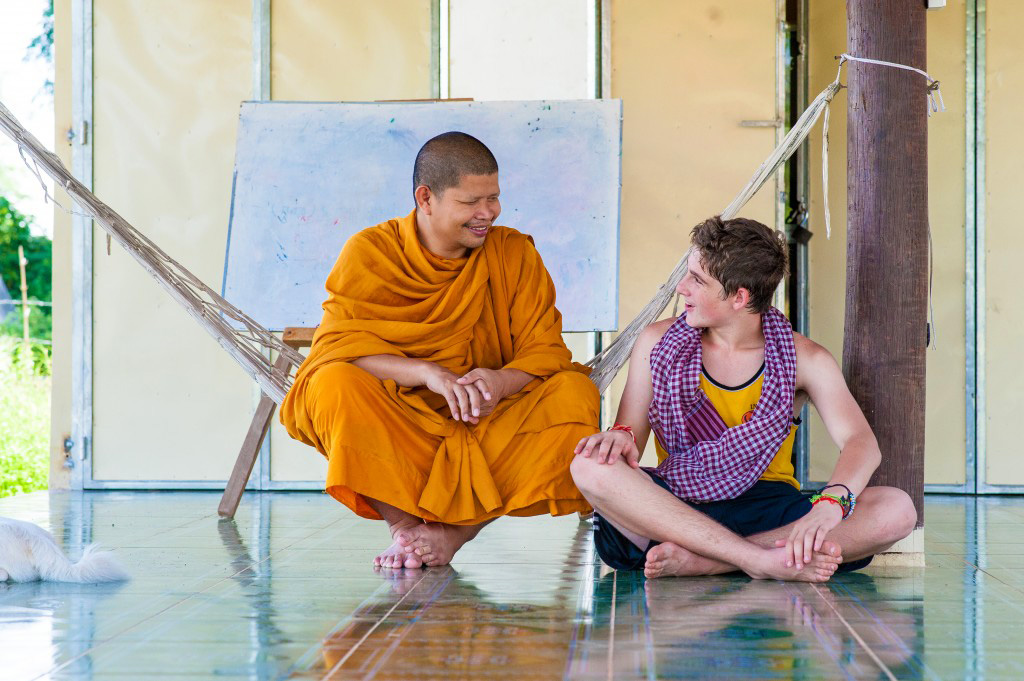 A student sits cross-legged sharing a conversation with a monk in orange robes