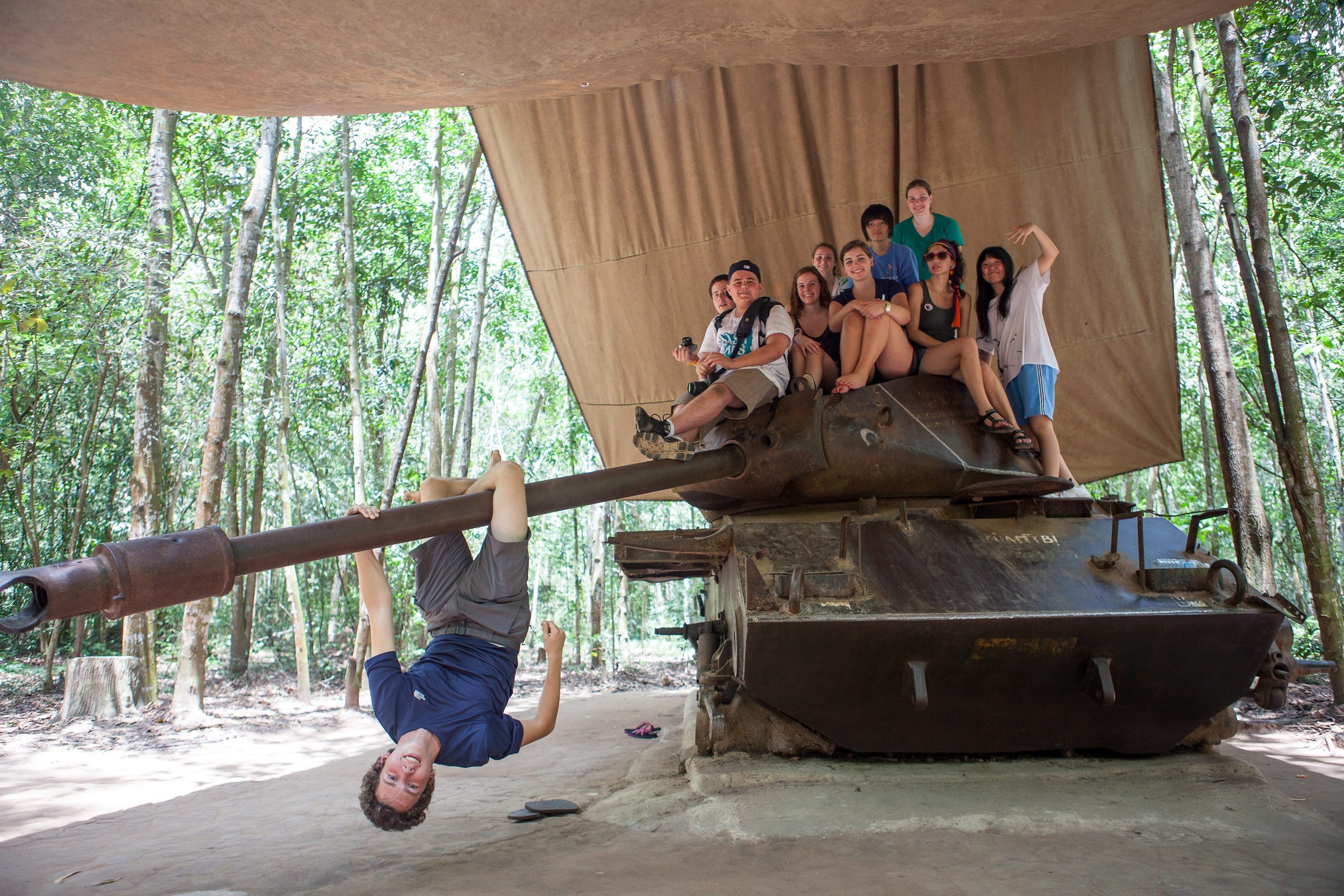 Students sit atop an old military tank under a canopy while one hangs upside down from the gun barrel