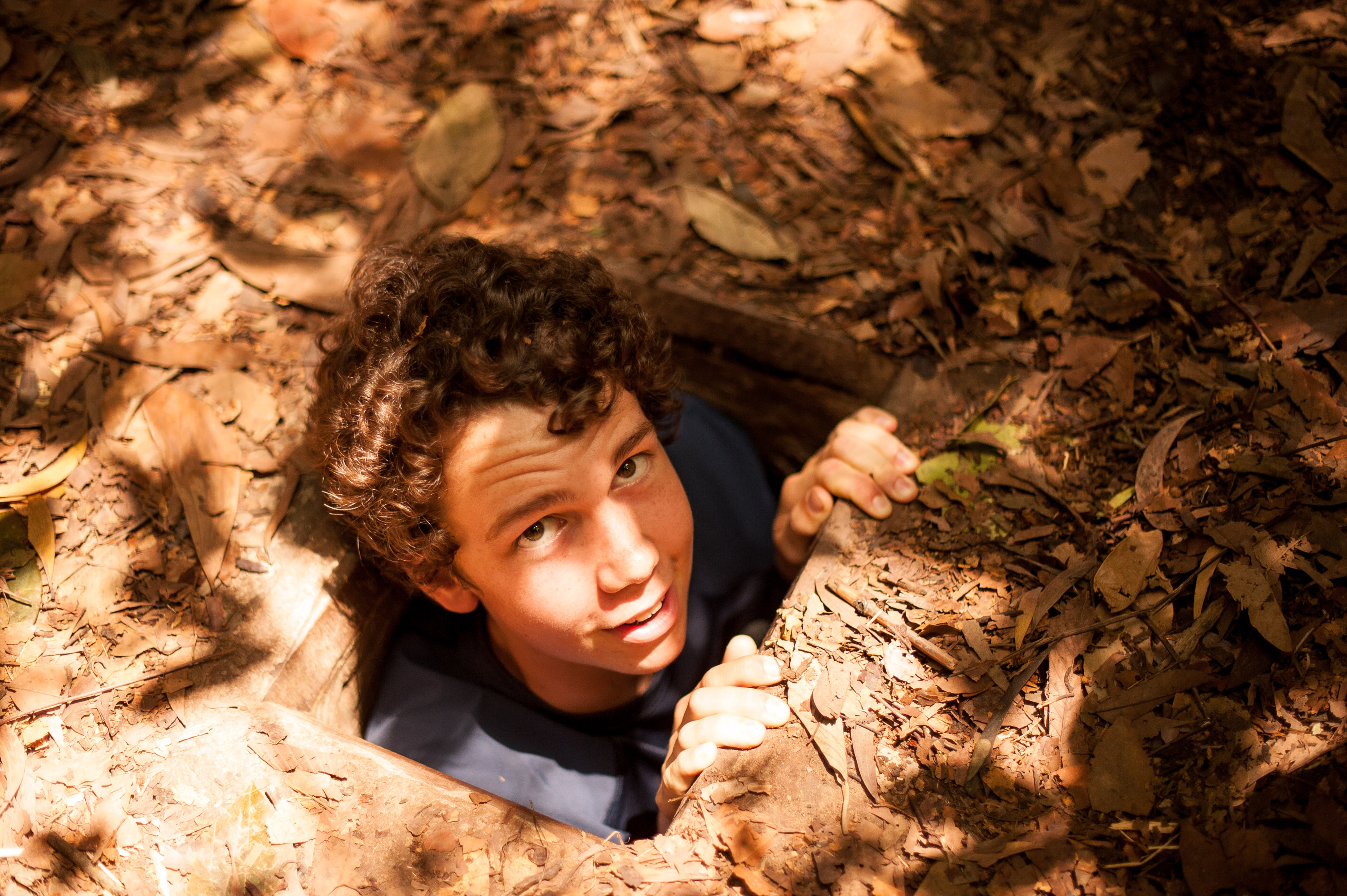 A student climbs out of a narrow tunnel opening covered in dry leaves