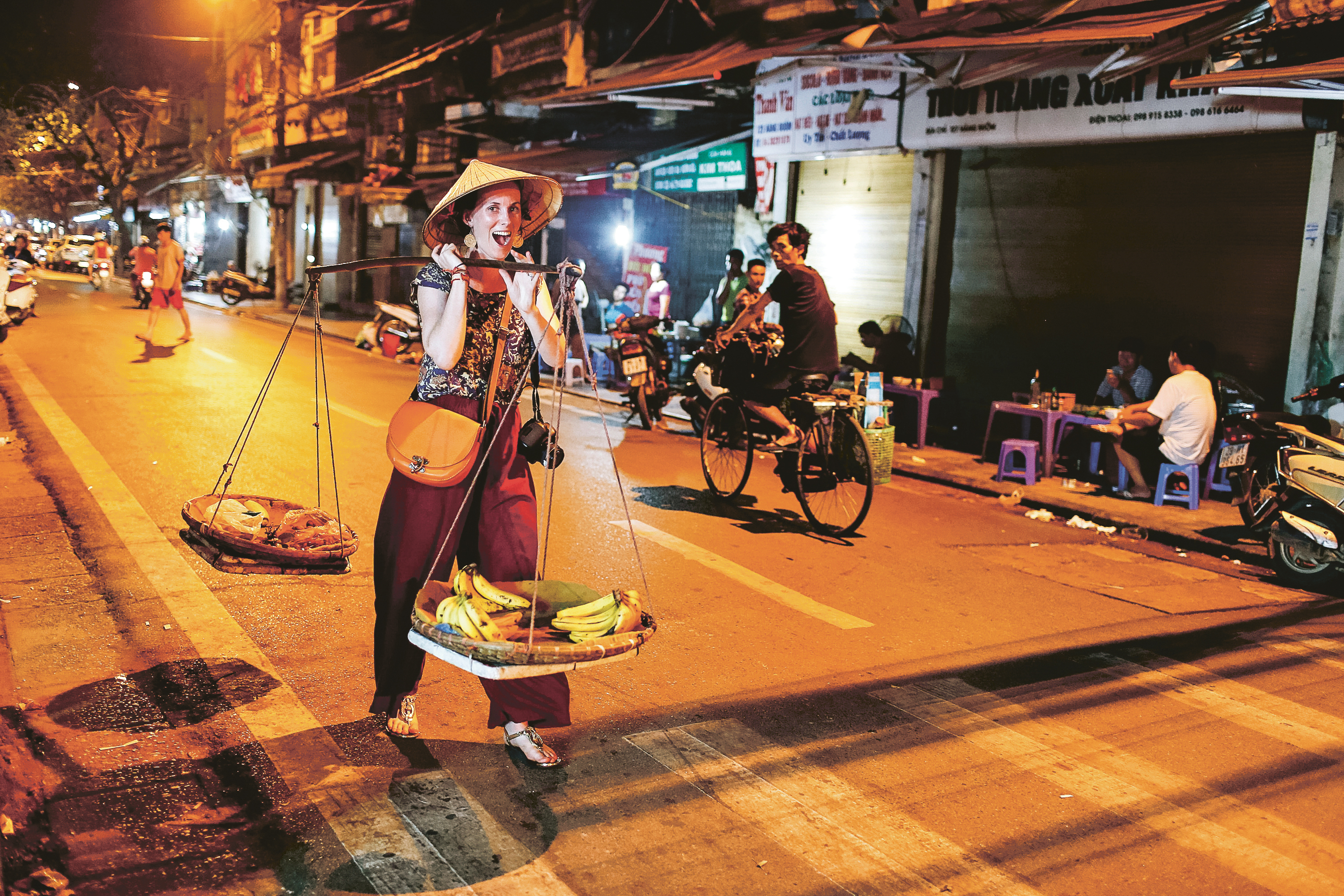 Student travelers watch a street vendor carry baskets of bananas on a shoulder pole through a Hanoi street at night