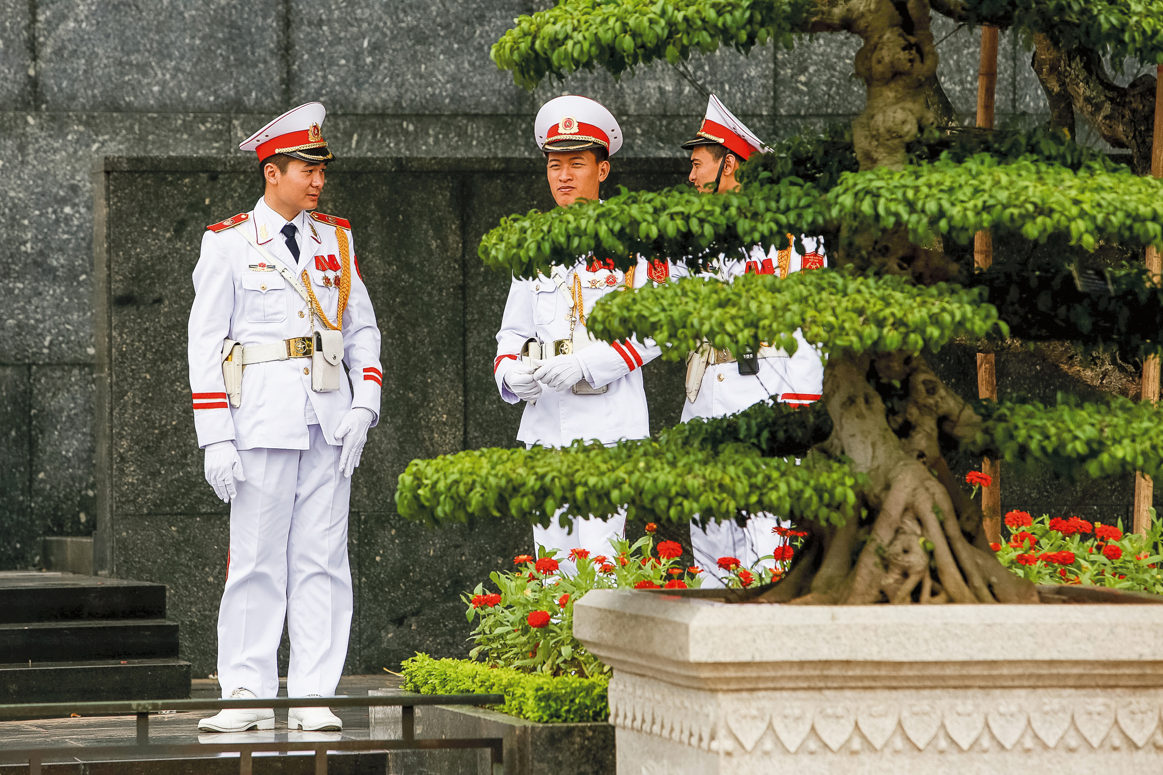 A school trip stops near a Hanoi monument where ceremonial guards in white uniforms stand by a trimmed tree