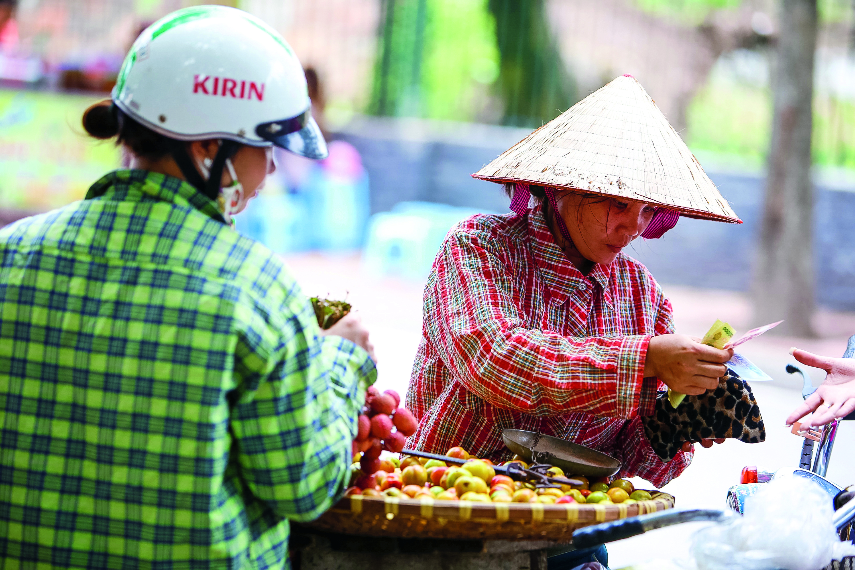 A vendor in a conical hat sells fruit from a street cart to a passerby