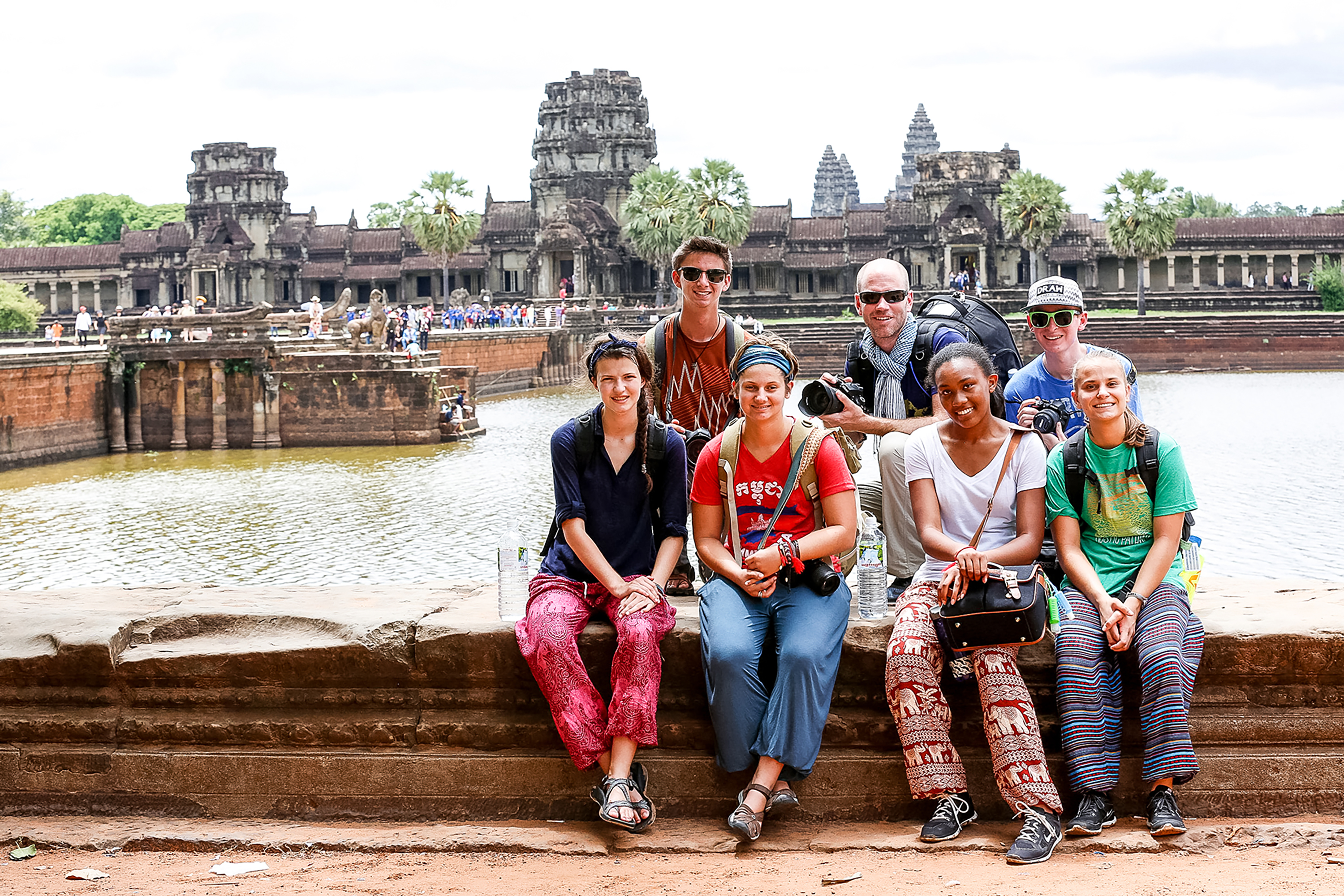 Students sit along a stone wall in front of Angkor Wat's moat and towers