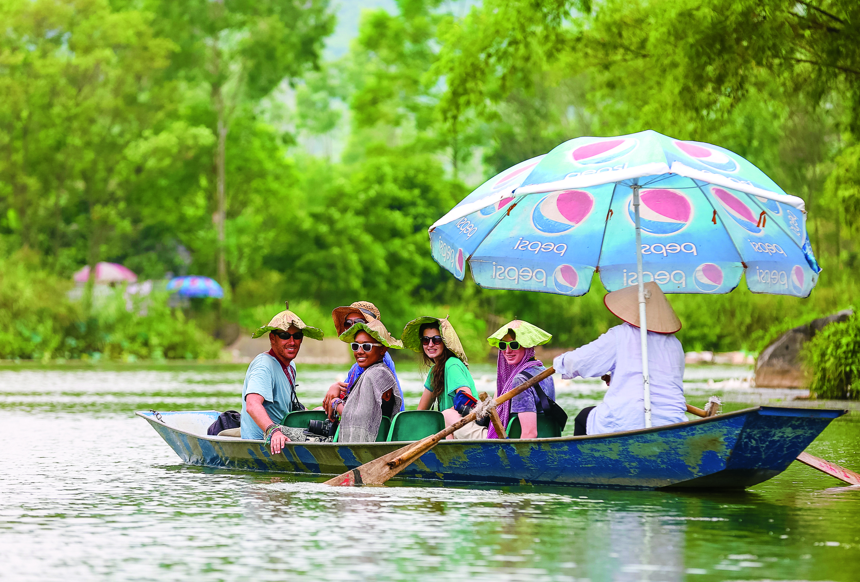 Students wear conical hats aboard a wooden riverboat under a large umbrella