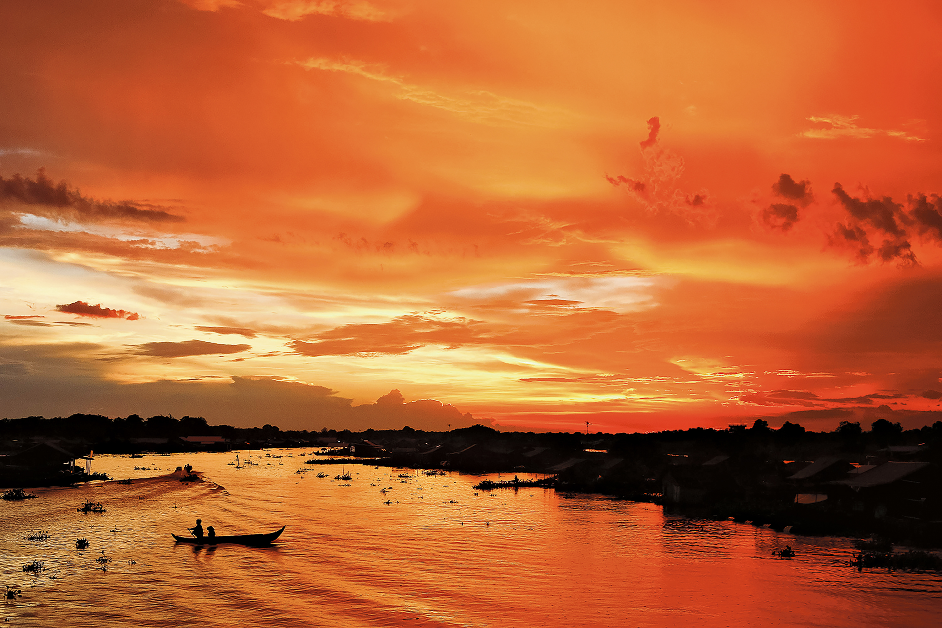 Teens cross a river by small boat under a vivid orange sunset near a floating village