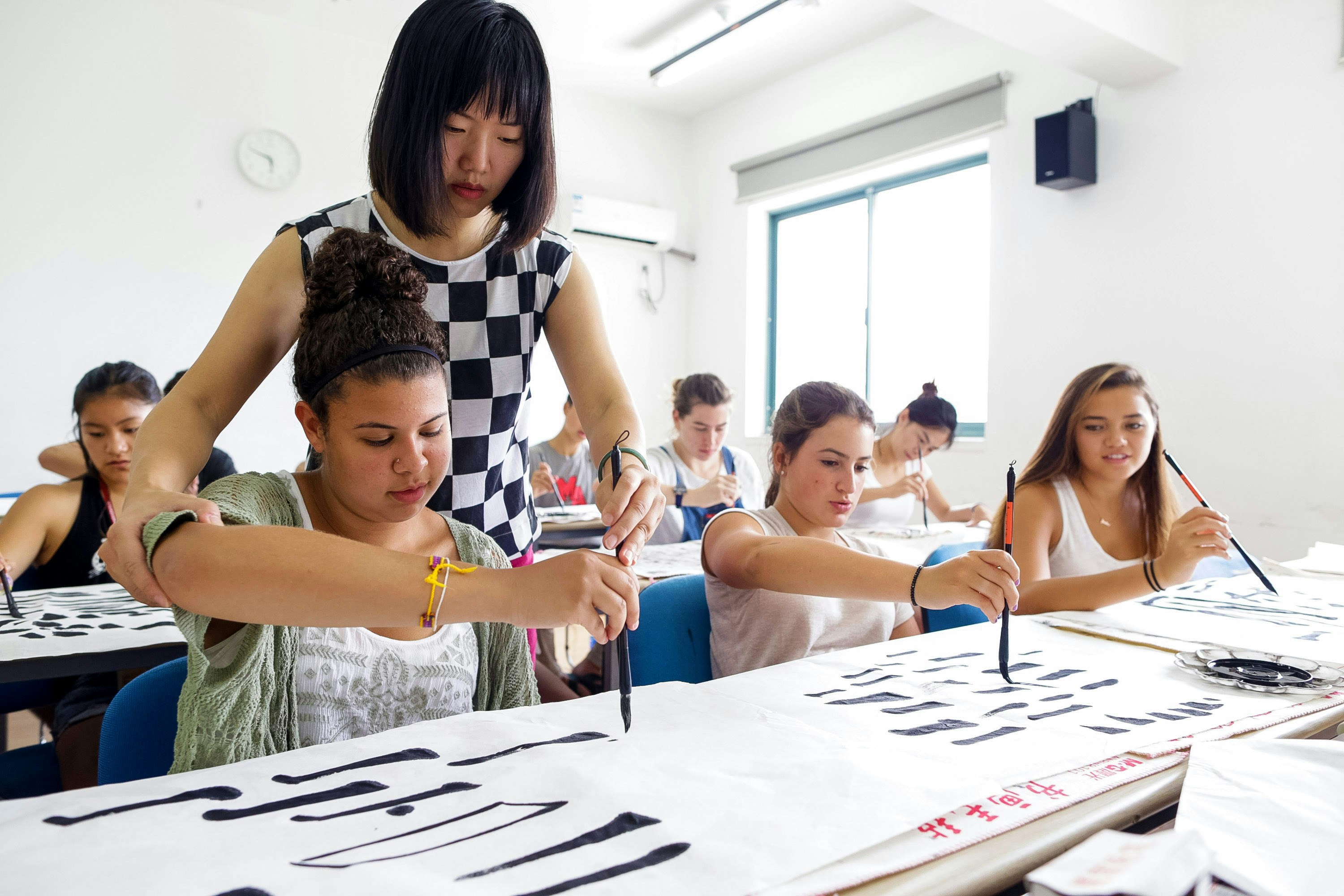 A Chinese teacher helping guide students in a classroom working on Chinese calligraphy.