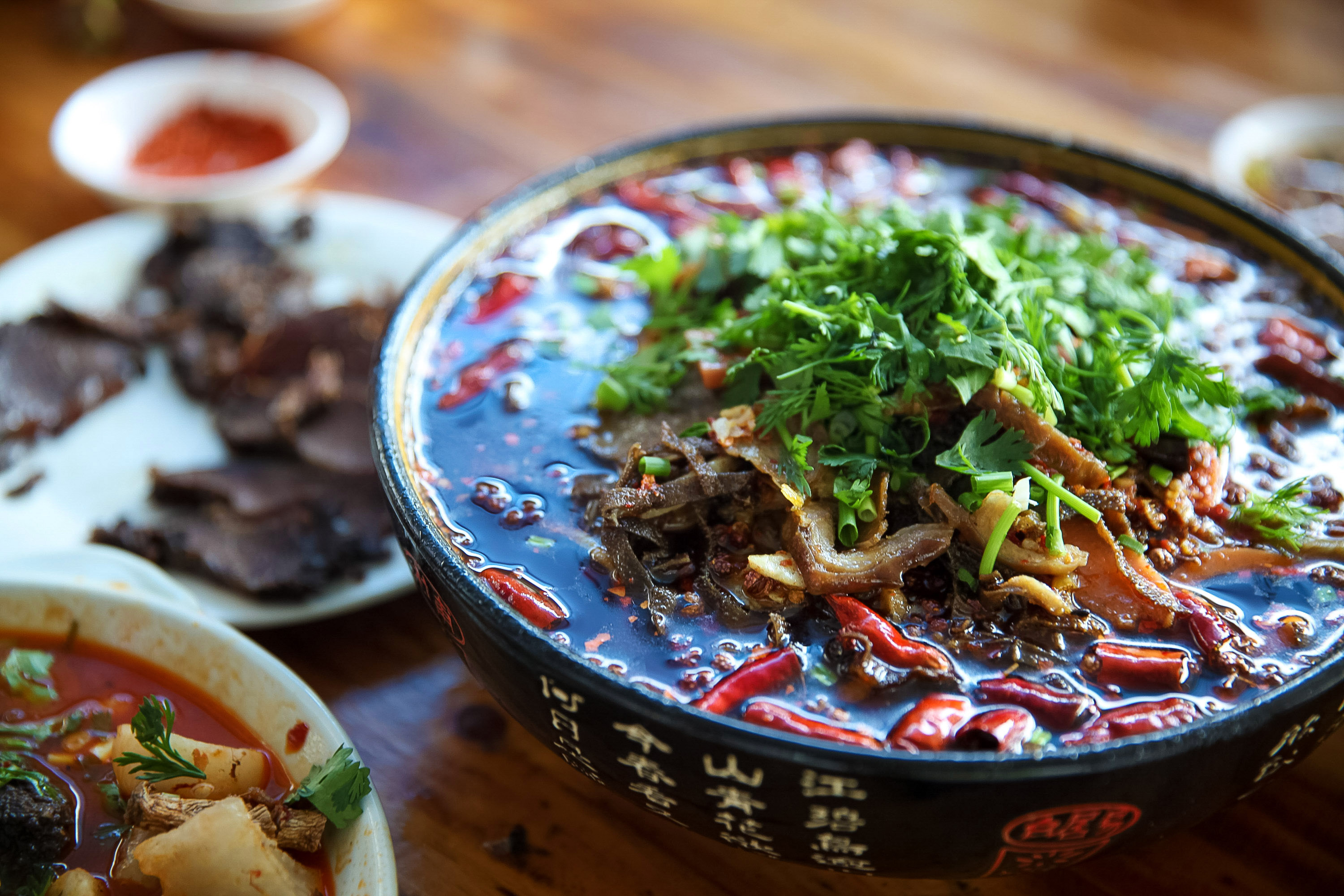 A bowl of spicy Sichuan noodles topped with cilantro and chili peppers, served alongside sliced meat and side dishes