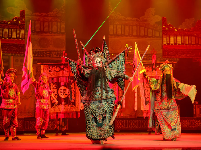 A performer in embroidered armour with banners rising from the shoulders holds a pose under red stage light.