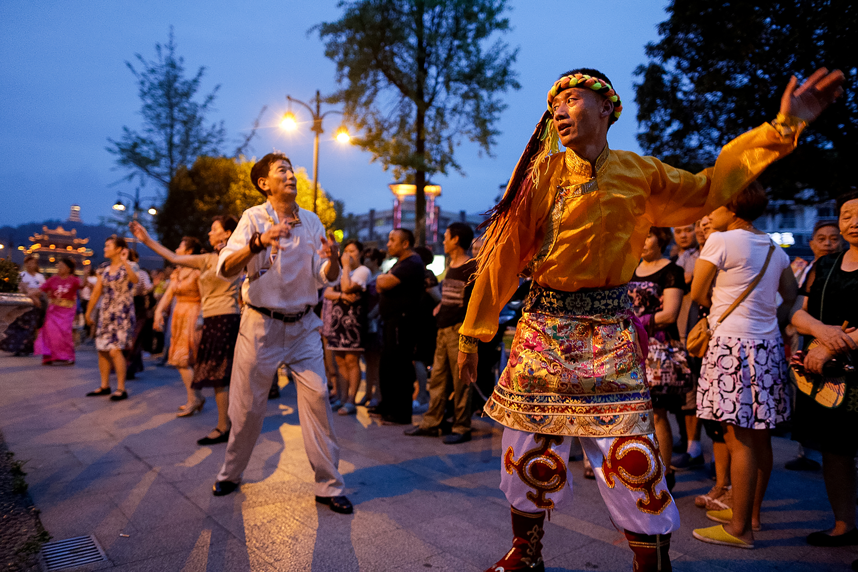 A performer in traditional dress dances at dusk as onlookers gather along a street