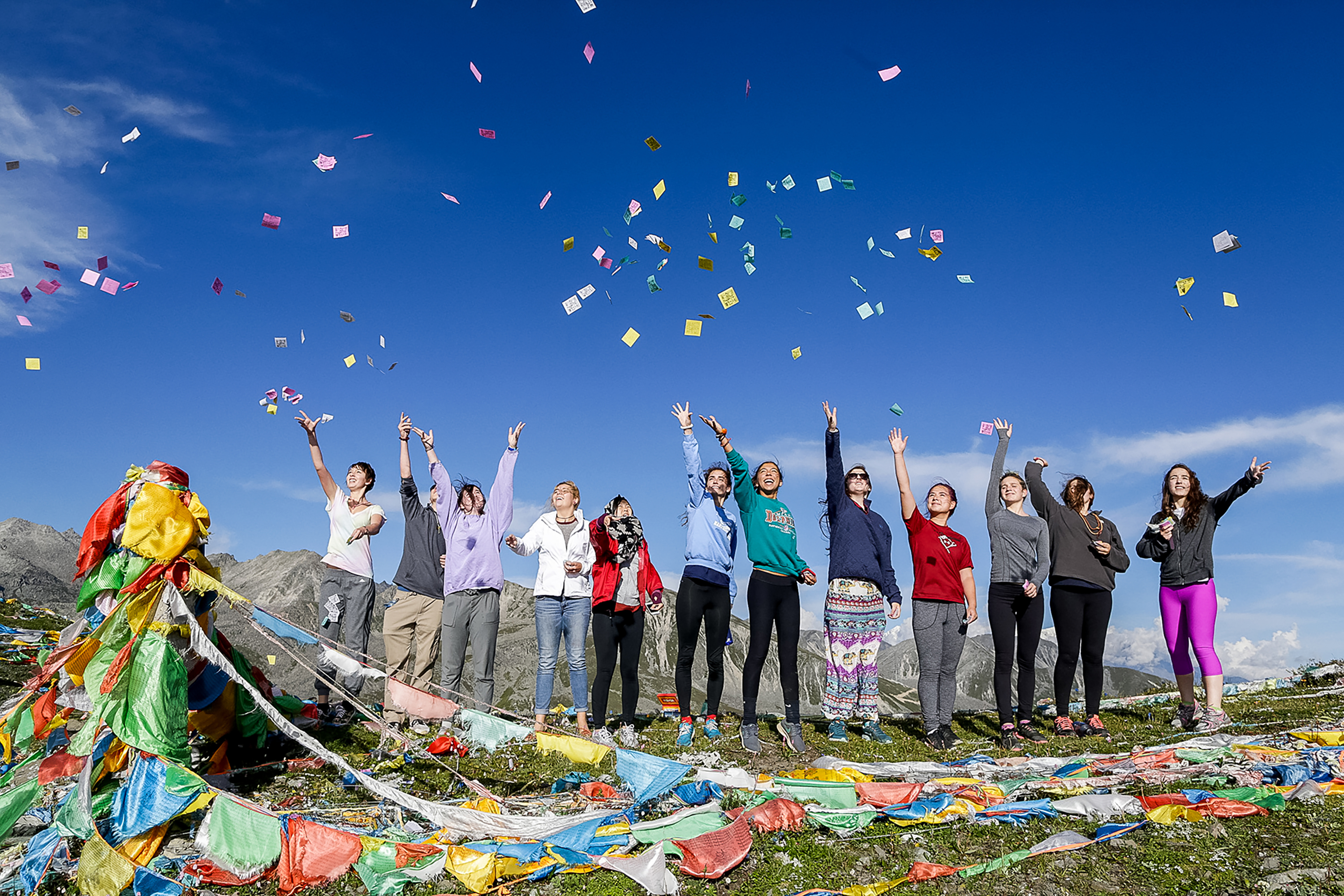 Students raise their arms as colorful paper flies through the air on a mountaintop strung with prayer flags