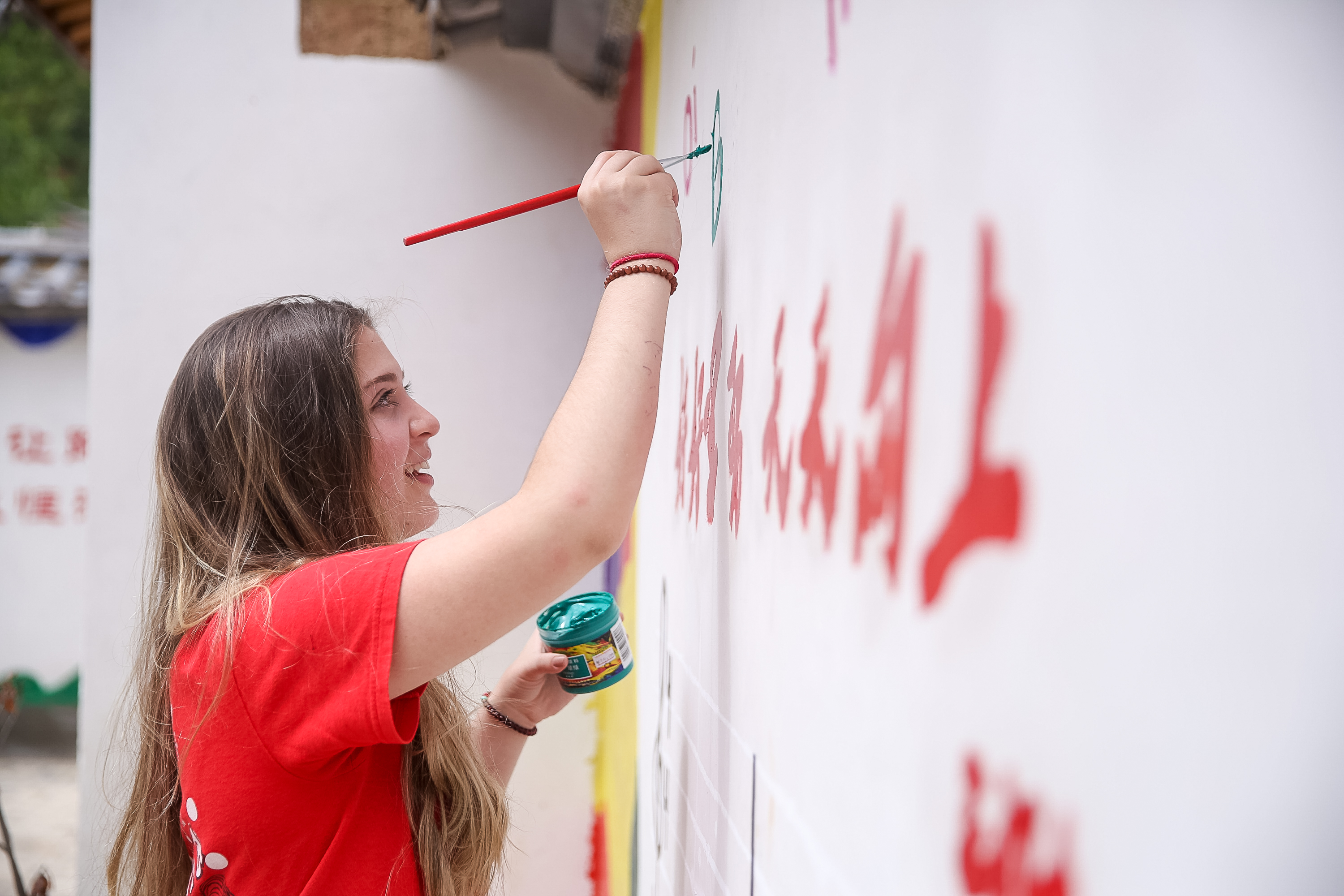 A student in a red T-shirt paints large red Chinese characters onto a white wall with a long brush.