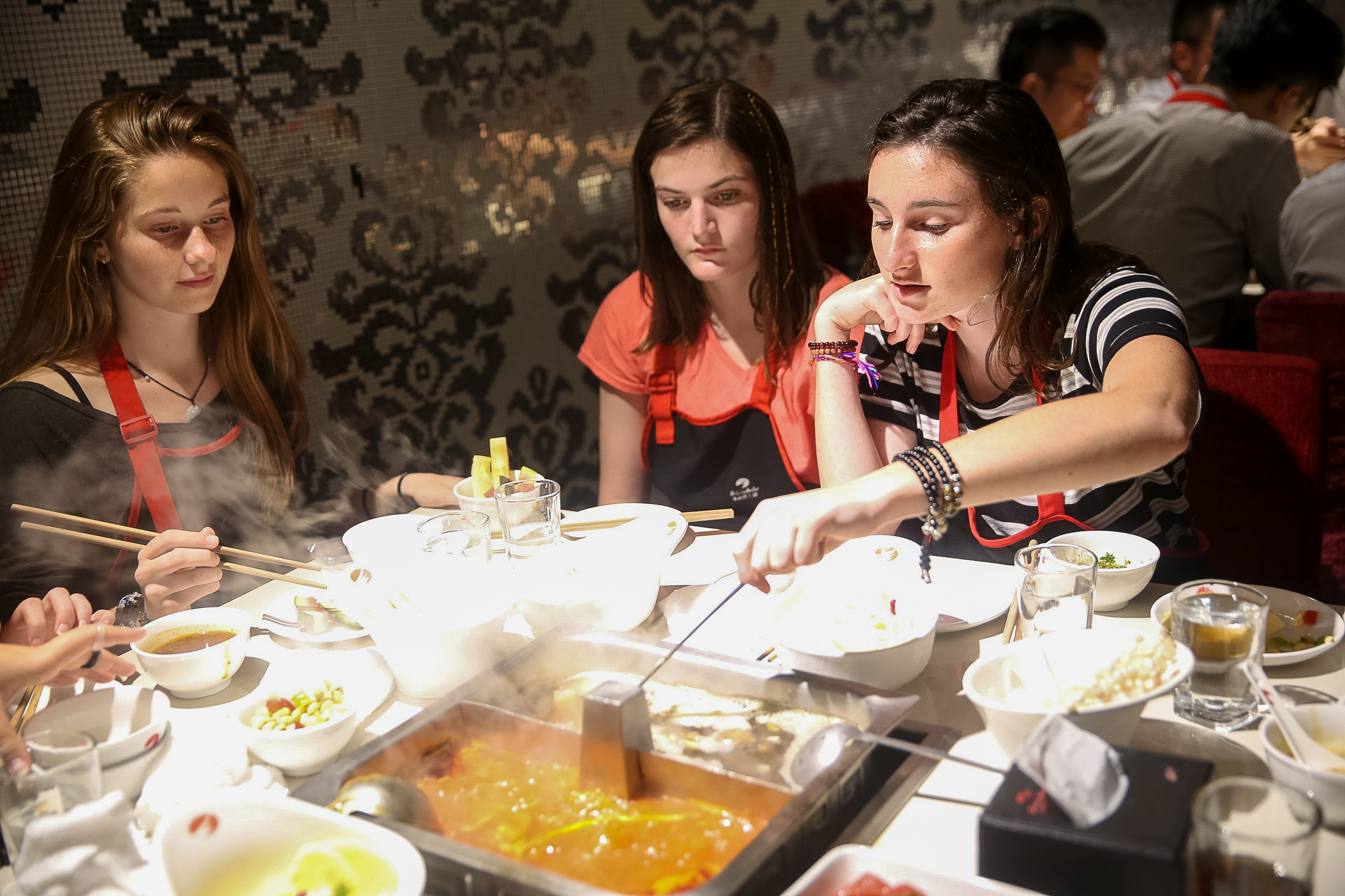 Two students lean over a divided hotpot on a restaurant table, lifting food from the simmering broth with chopsticks.