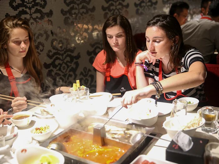 Two students lean over a divided hotpot on a restaurant table, lifting food from the simmering broth with chopsticks.