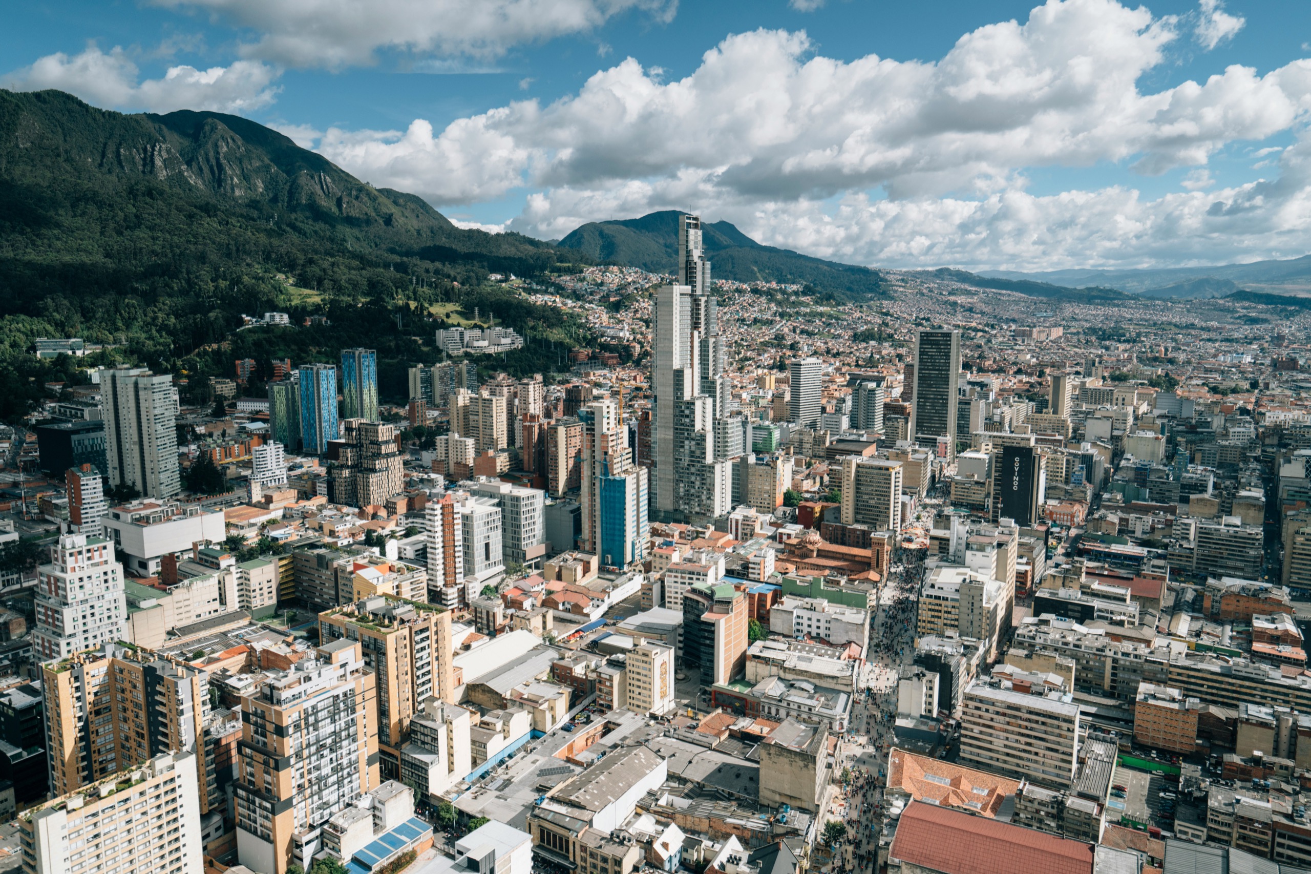 Bogotá's high-rise downtown in Colombia spreads across the valley floor beneath green Andean peaks under a partly cloudy sky.