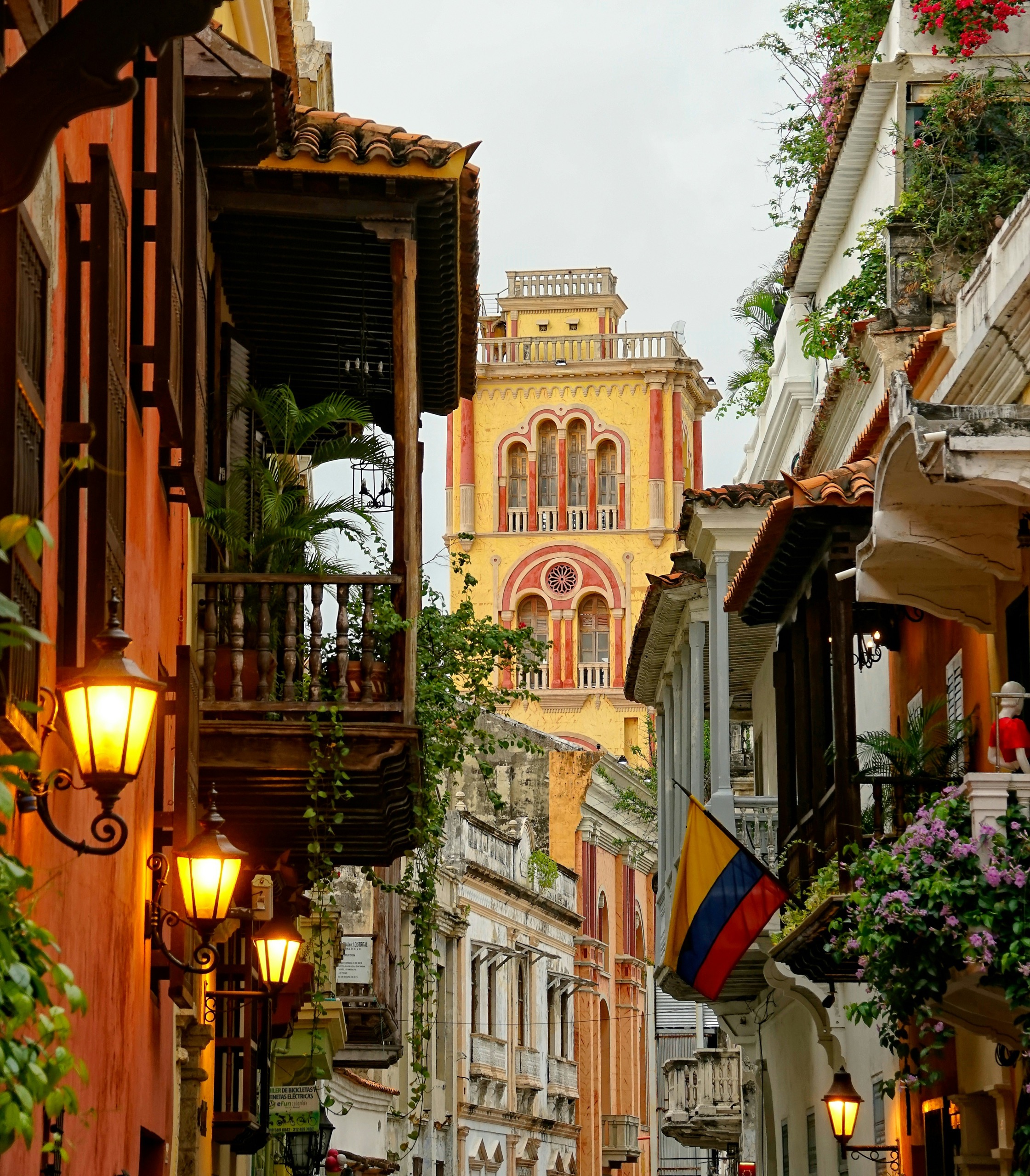 Balconies and a colonial bell tower rise above a narrow street in Cartagena's walled city, string lights lit at dusk.