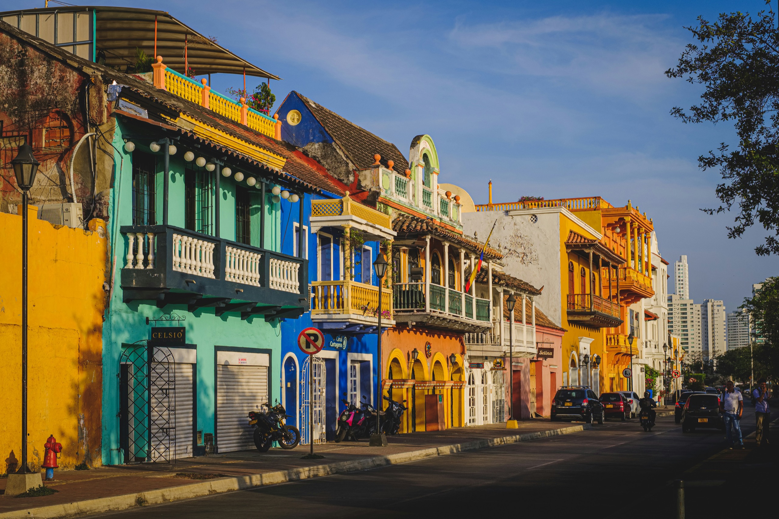 Two-story houses in turquoise, blue and gold line a sunlit street in Cartagena's Getsemaní neighborhood.