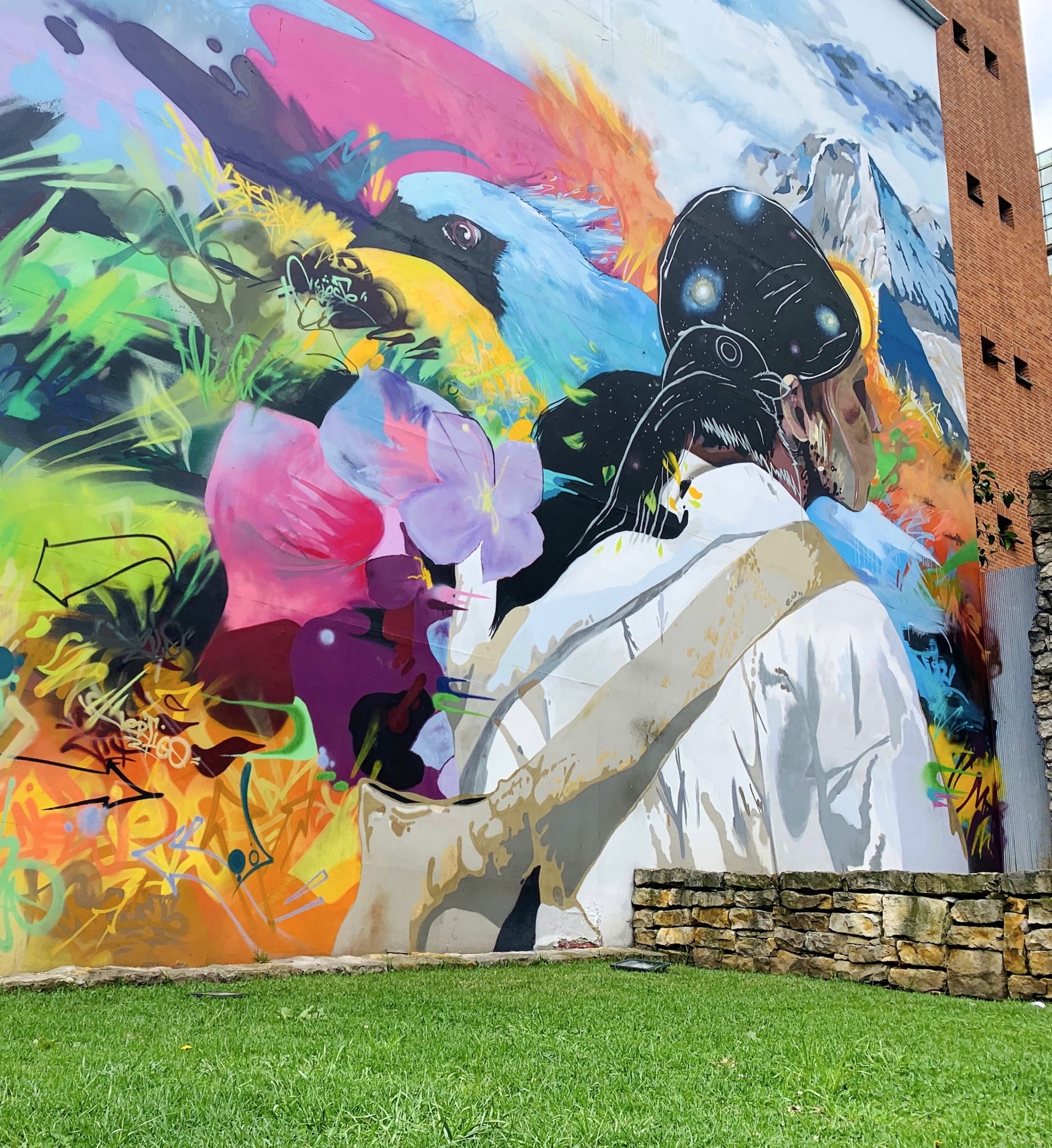 A street mural in Bogotá: a figure in white beside a bird, flowers and snow-capped mountains, painted across a building wall above a stone terrace.