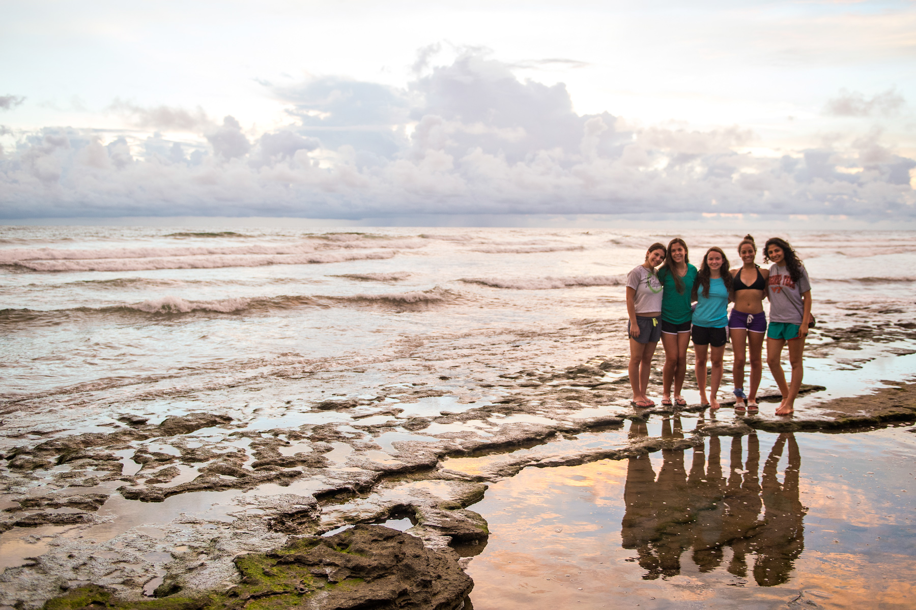 Students stand together on a tidal rock ledge as waves roll in at sunset