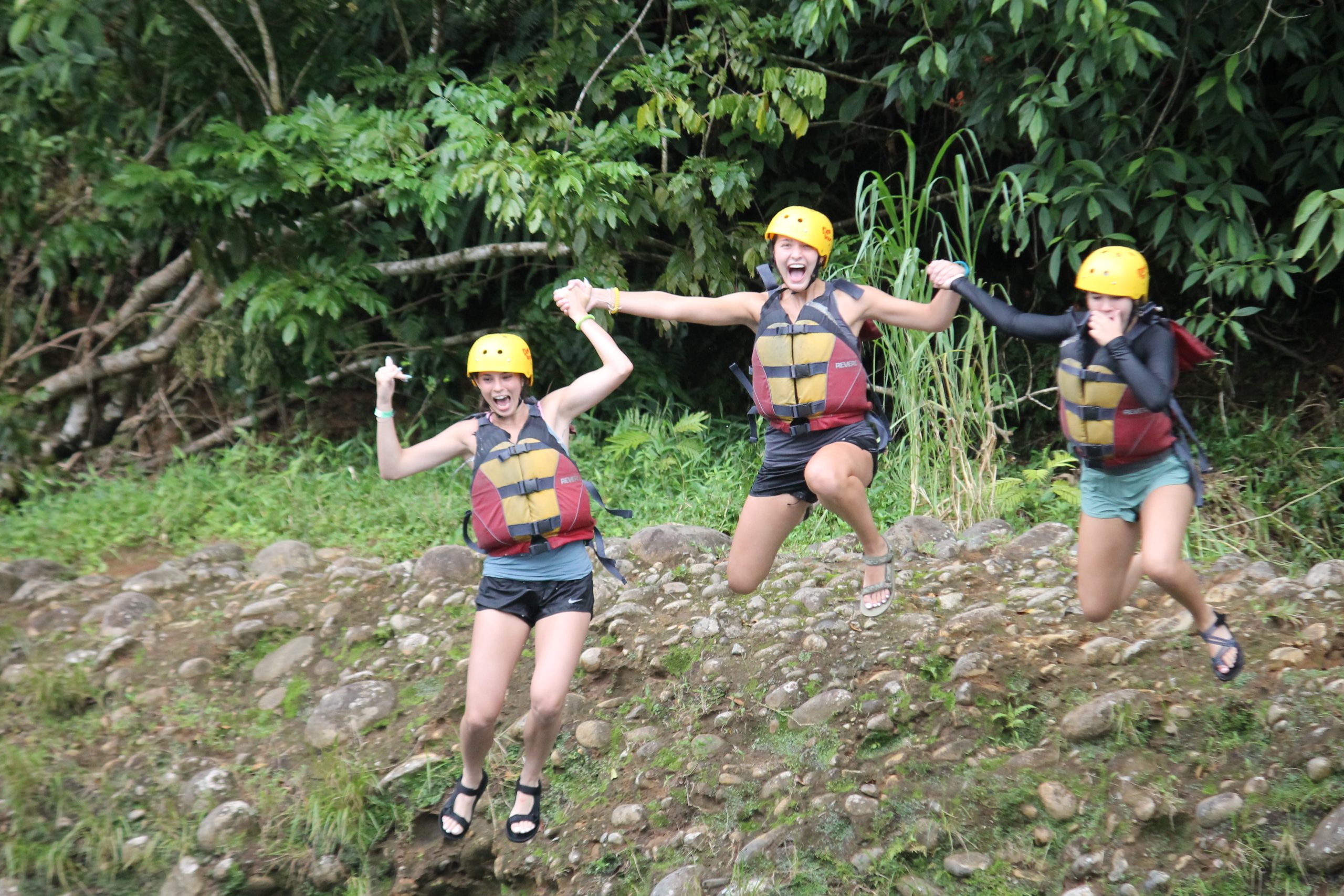 Students in helmets and life jackets jump for joy on a rocky riverbank in Costa Rica
