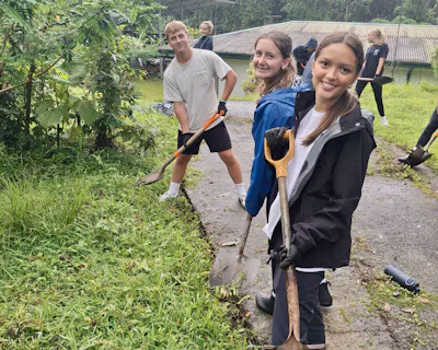 Three students pause with shovels on a wet path, clearing ground beside a field station