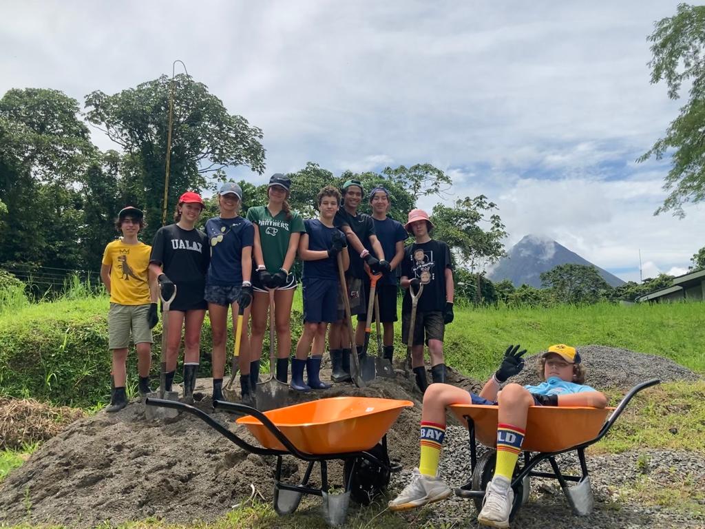 Students pose with shovels beside wheelbarrows on a service site, a volcano rising in the distance