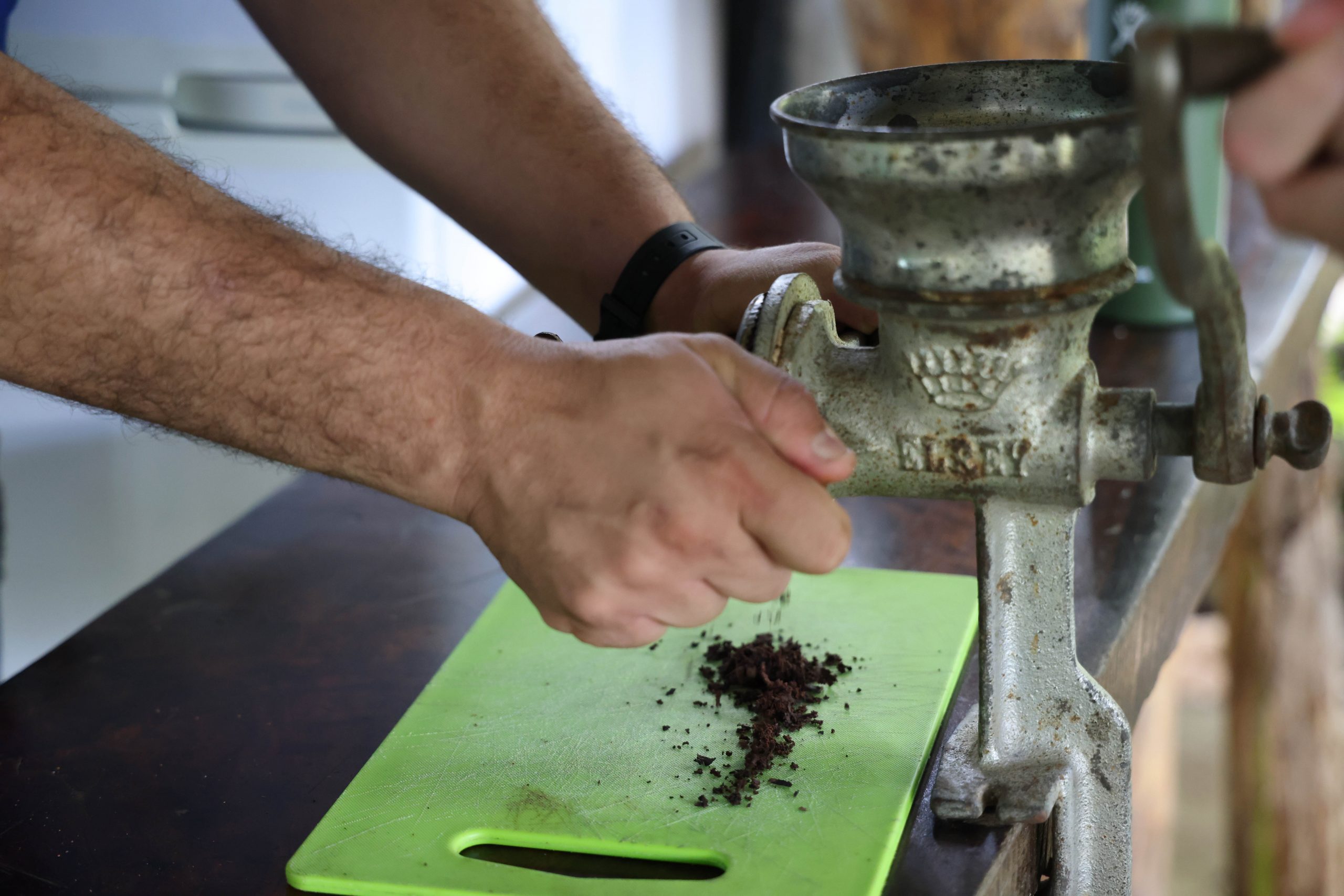A hand-crank grinder processes cacao onto a green cutting board