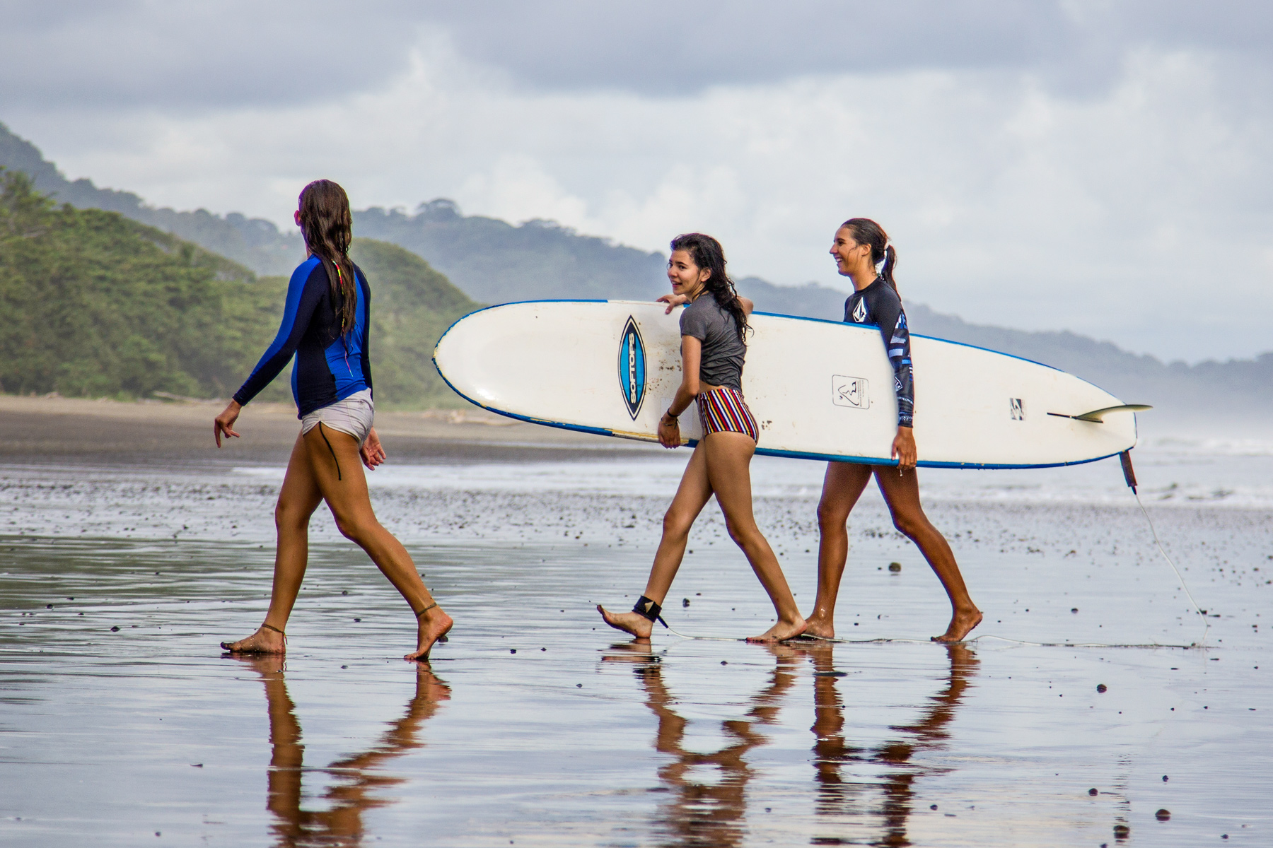Students carry a surfboard while walking along a wet beach in Costa Rica
