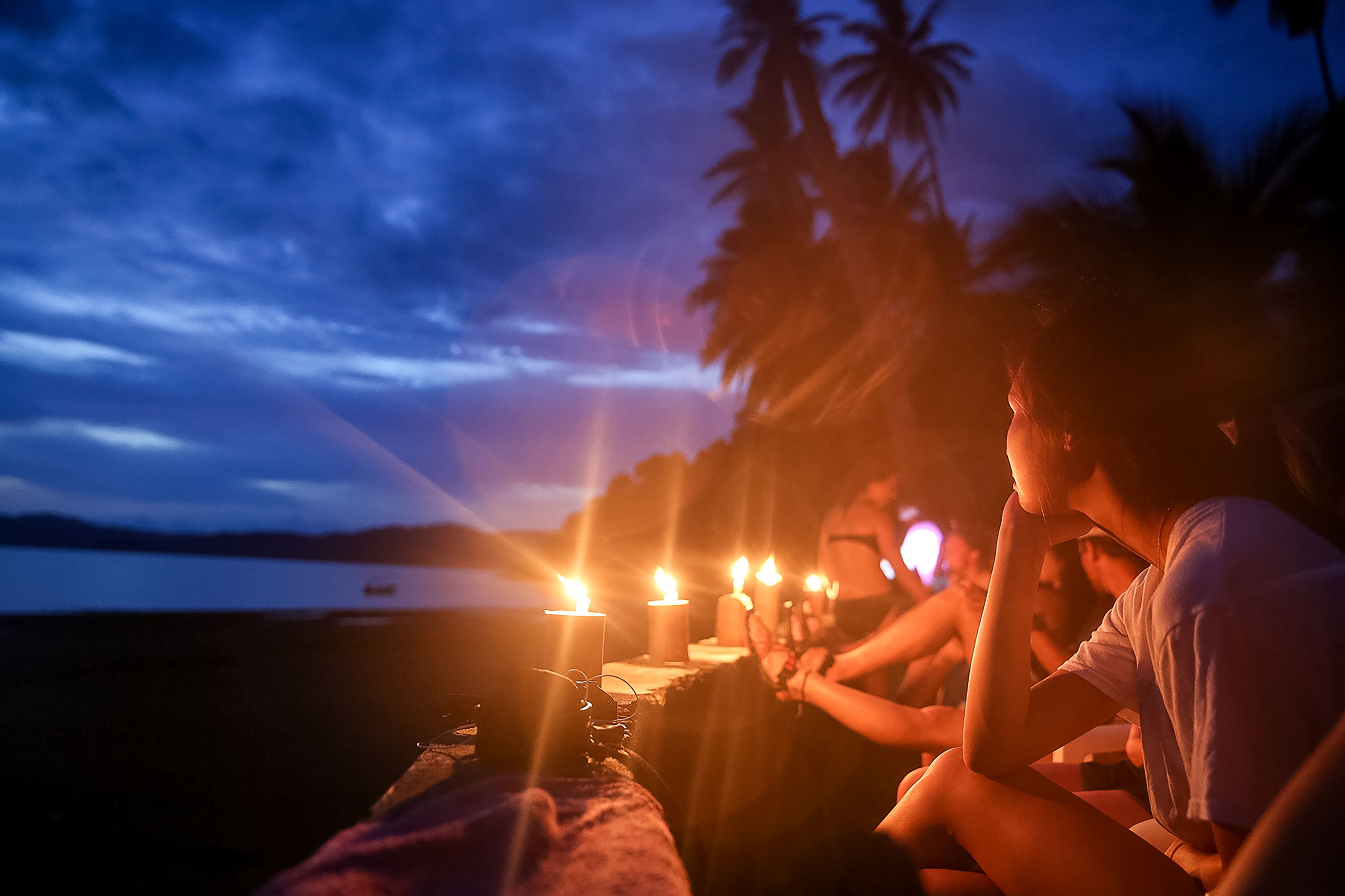 Students sit along a candlelit beach wall at dusk with palm trees silhouetted behind them