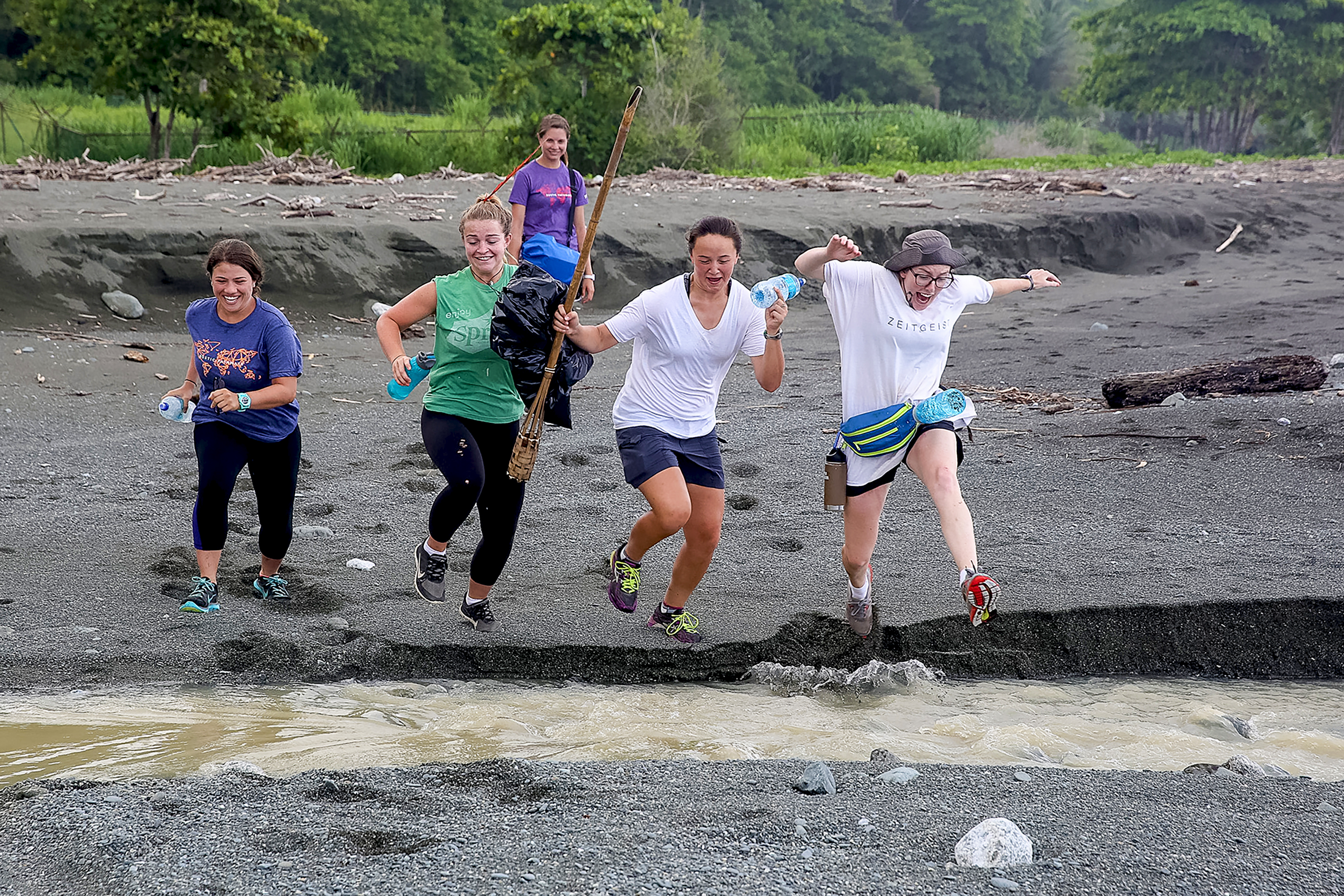Students jog along a muddy riverbank carrying gear and water bottles