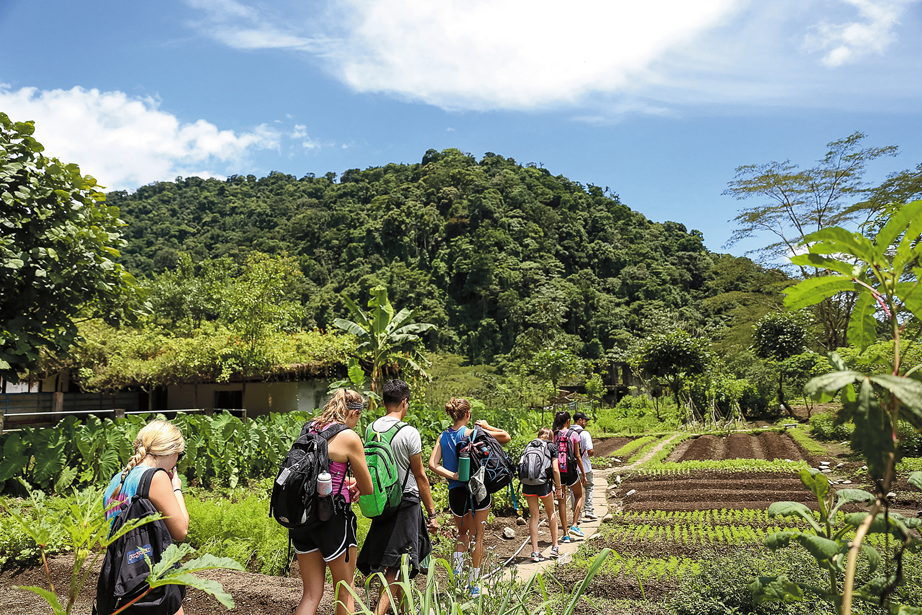 Students with backpacks walk a path beside farm fields with forested hills behind