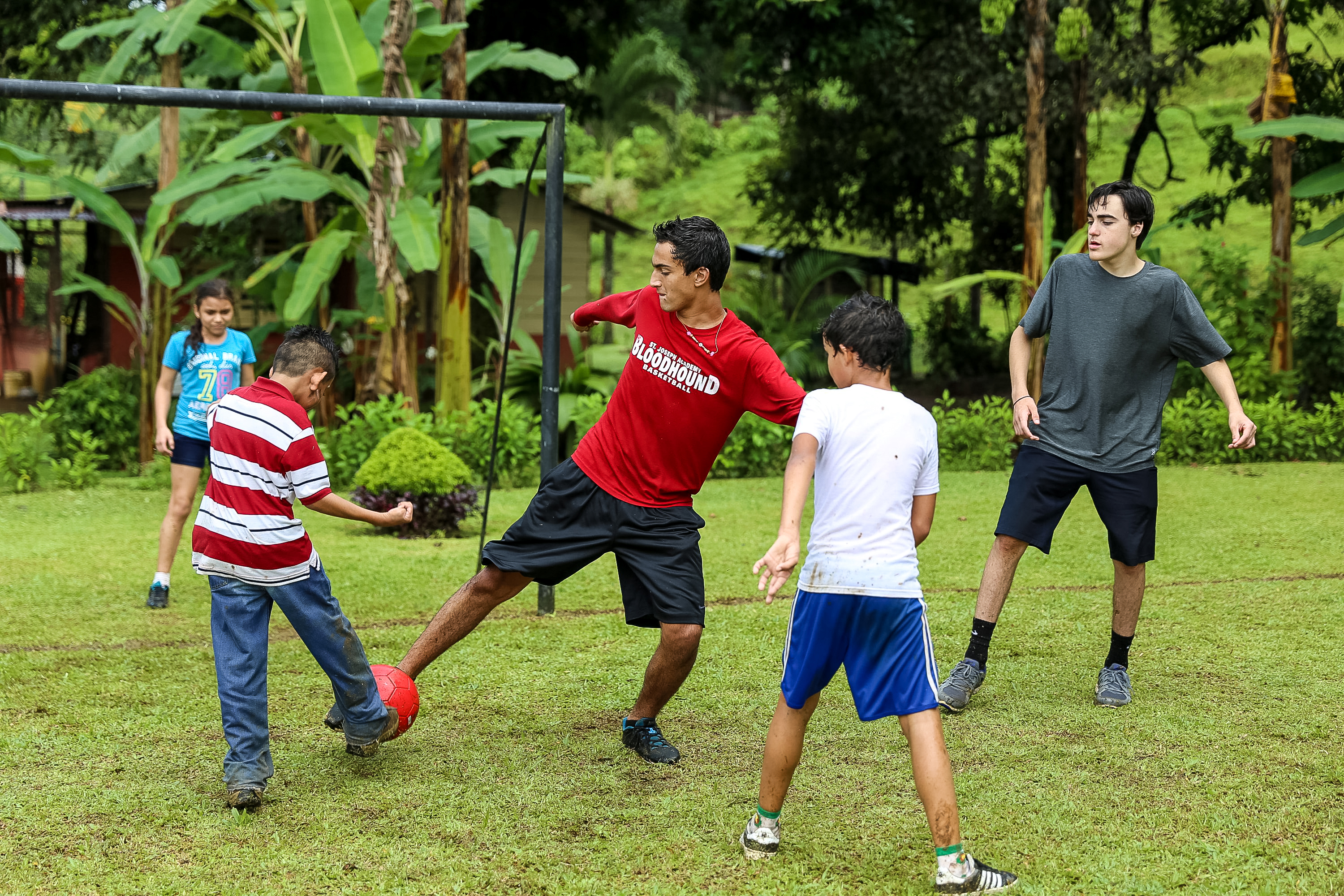 Students play soccer together on a grassy field surrounded by trees