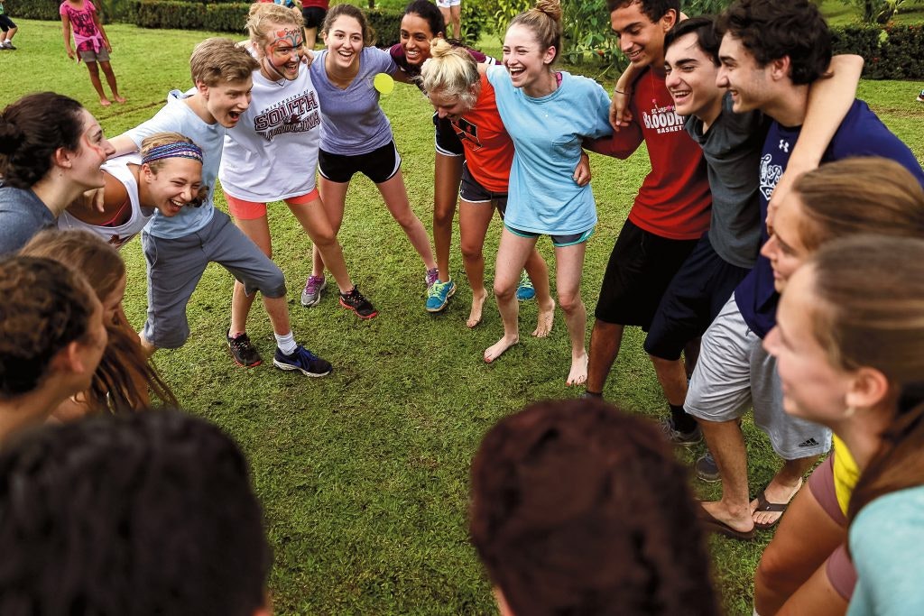 A group of smiling students stands in a circle outdoors, arms around each other, enjoying a fun and energetic moment together.