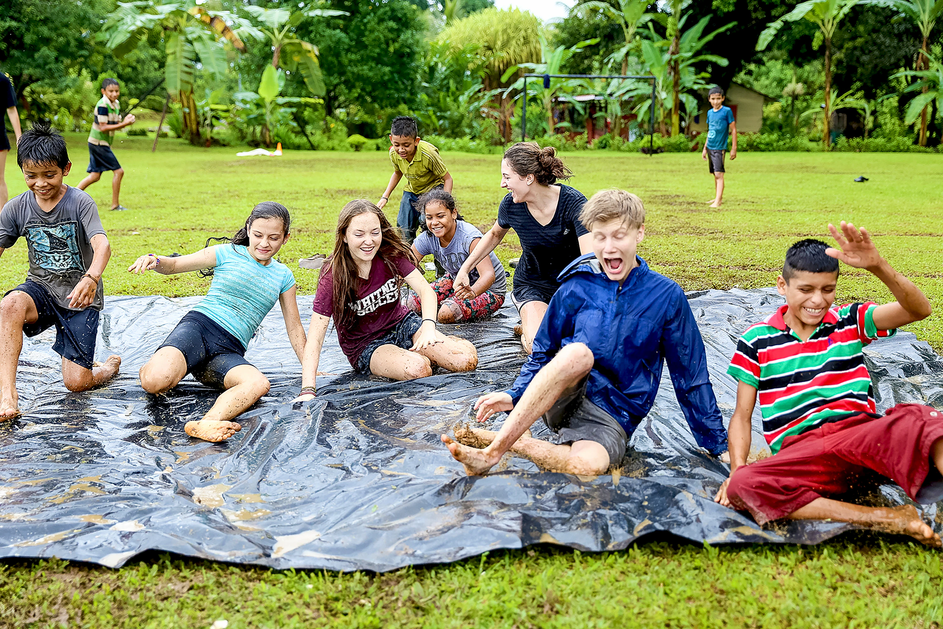 Teen travelers in Costa Rica slide and play with children on a muddy tarp during a rainy day game