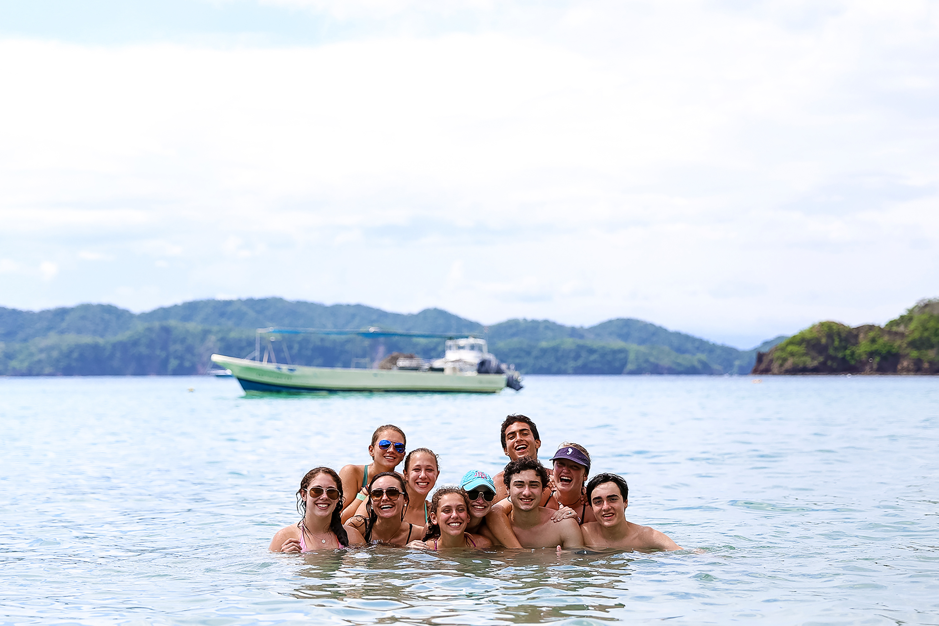 Students gather in the water for a group photo with a boat anchored near green hills behind them