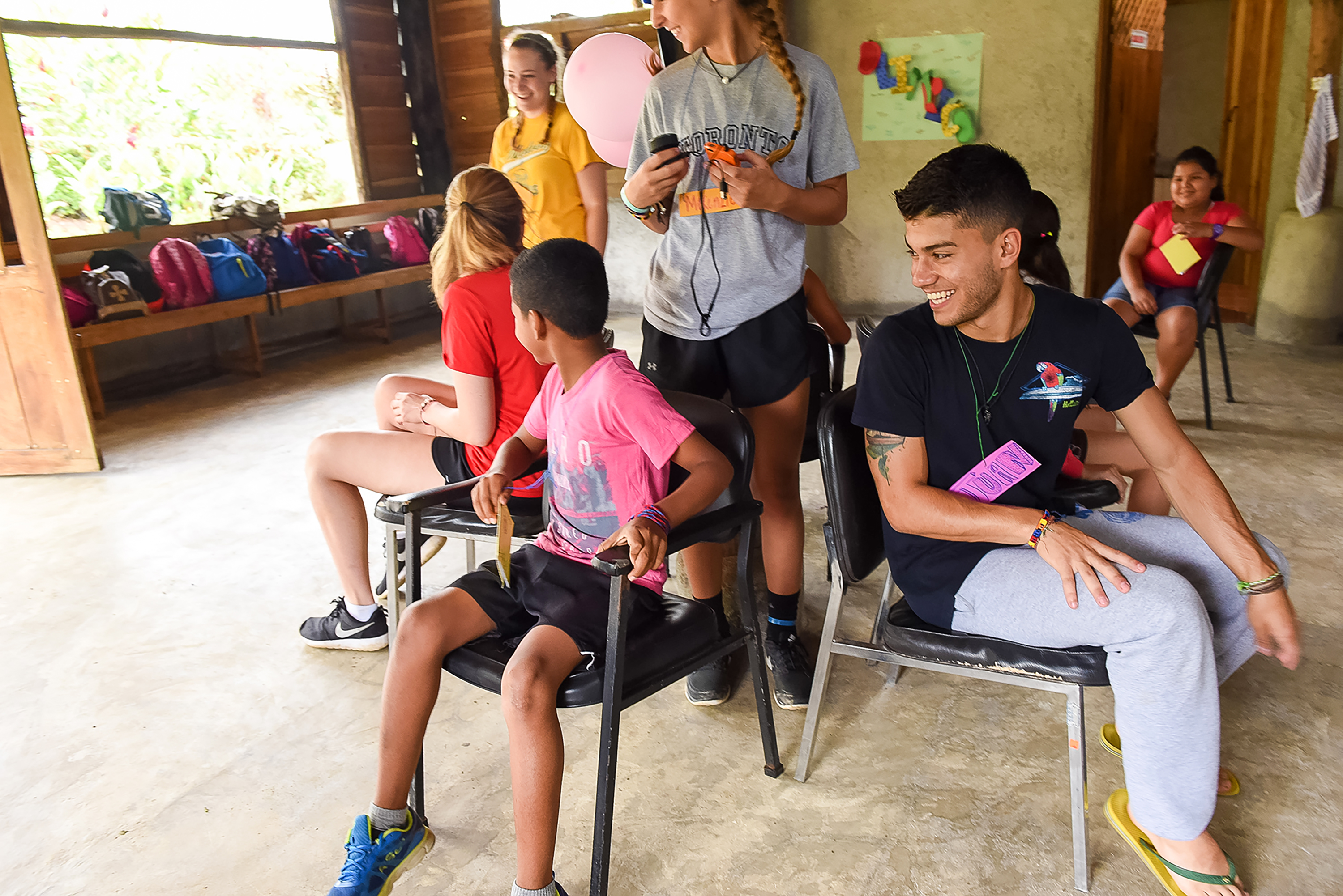 Teens sit in a circle of chairs, chatting with local children during a camp activity
