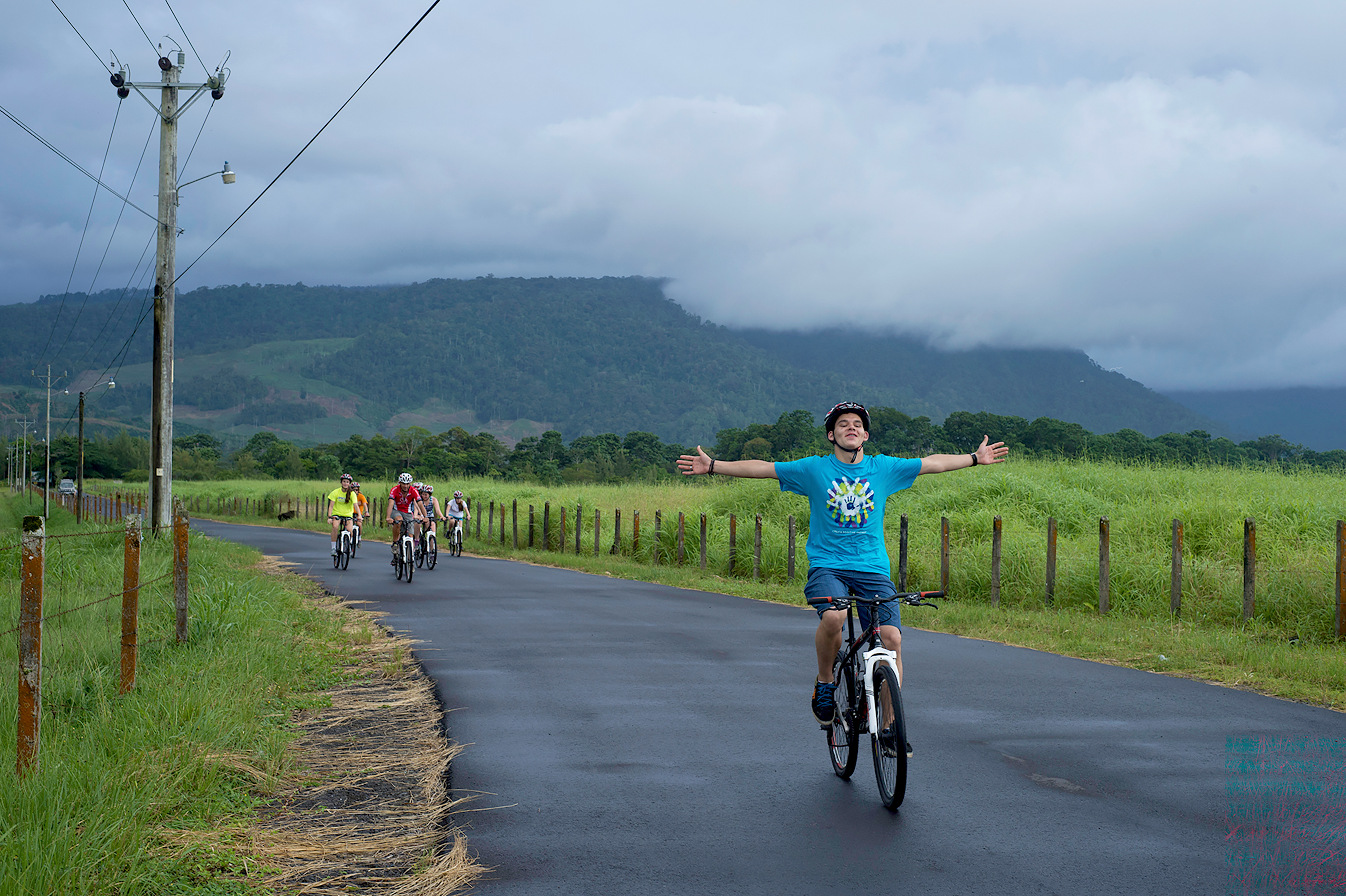 Student cycling with arms outstretched on a rural road, mountains behind