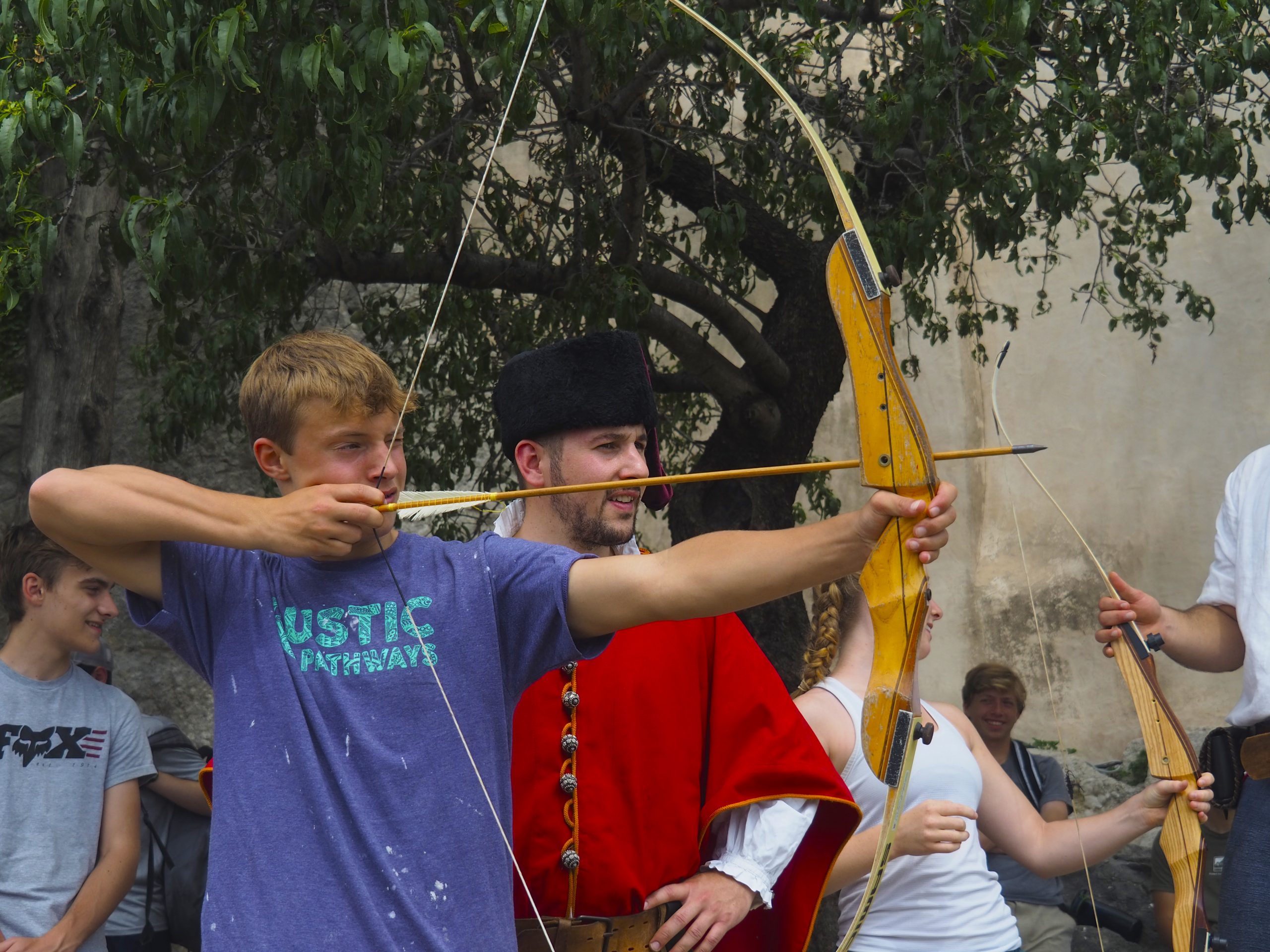 A student aims a traditional bow while a costumed guide helps steady his aim