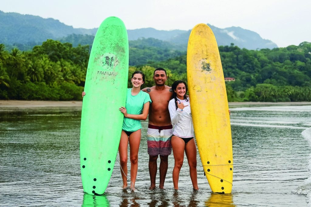 A surf instructor posing with two teen travelers holding surf boards on the beach