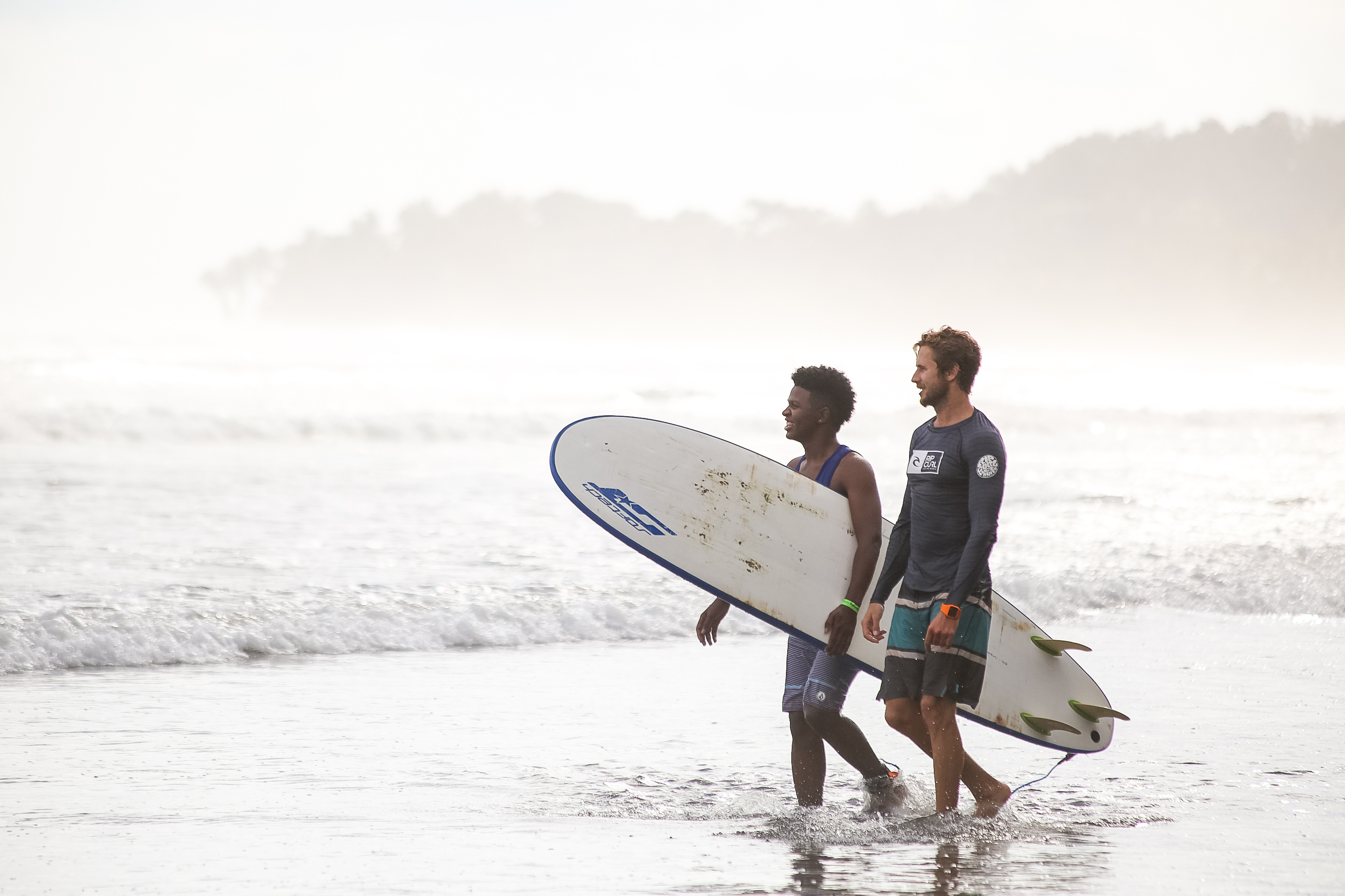 Teen travelers carry surfboards while walking along the shoreline